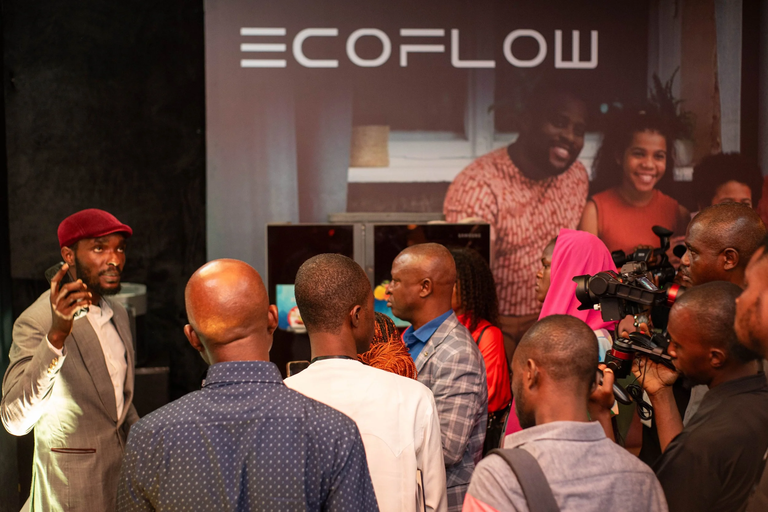 A large crowd of people attending an indoor event with a screen displaying the word 'ECOFLOW' and a smiling woman projected on the screen. The crowd includes a man with a red cap talking on a phone, and a photographer capturing the scene.