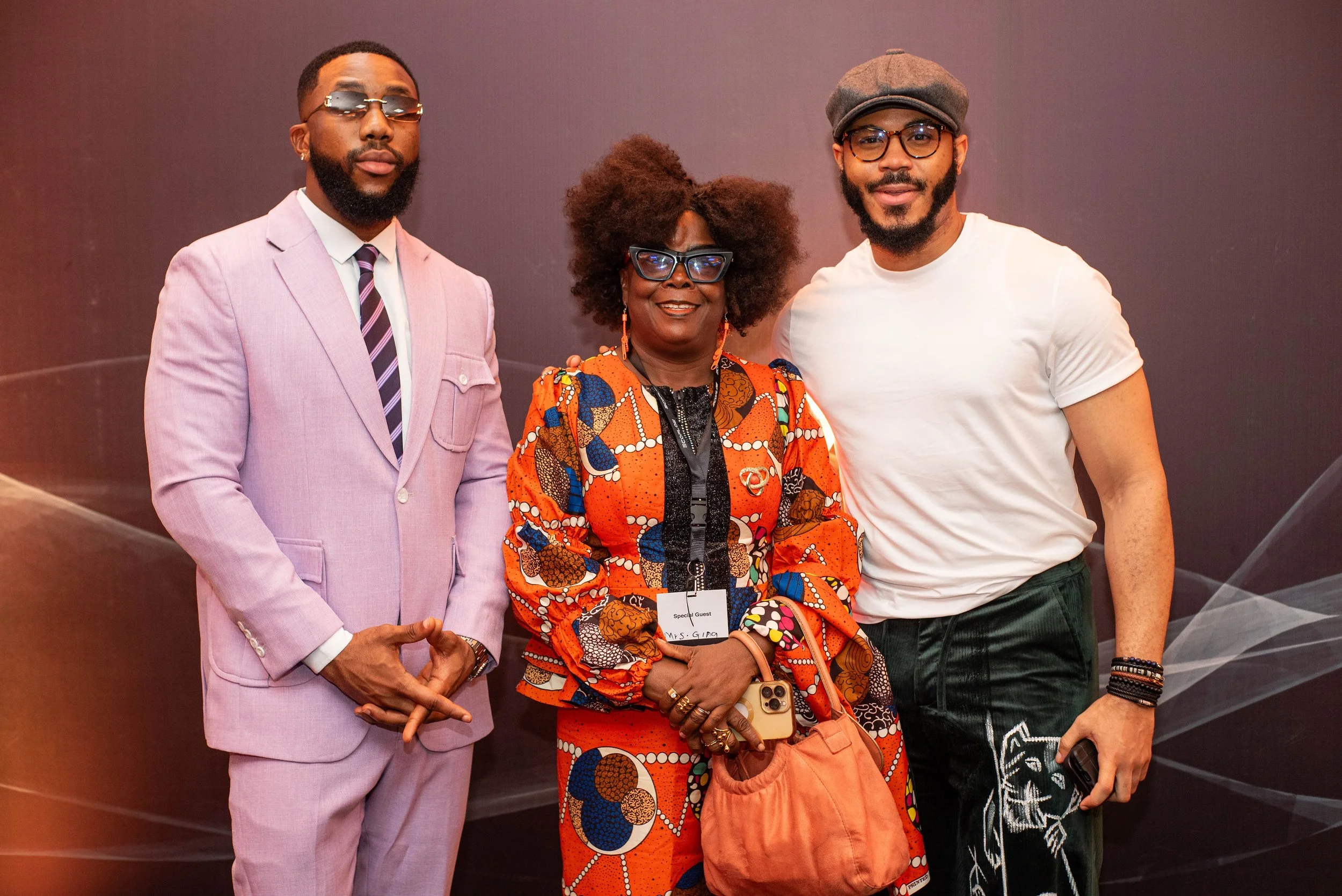 Three people stand together against a plain wall, smiling at the camera. The man on the left is dressed in a light pink suit with sunglasses, the woman in the middle wears colorful African print clothing and large glasses, and the man on the right we
