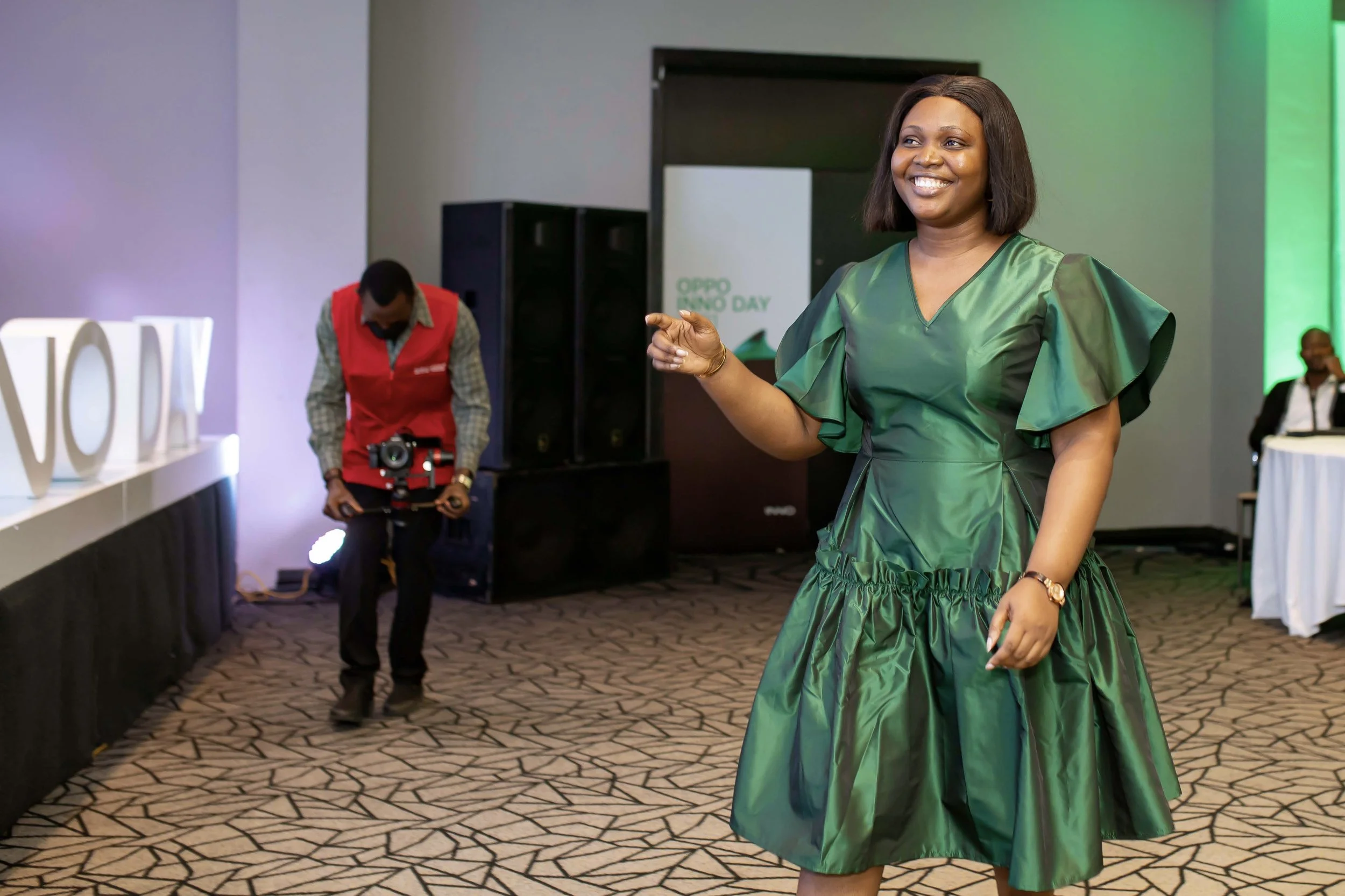 A woman in a shiny green dress smiling at an event, with people and a sign in the background that reads 'OPPO INNO DAY'.