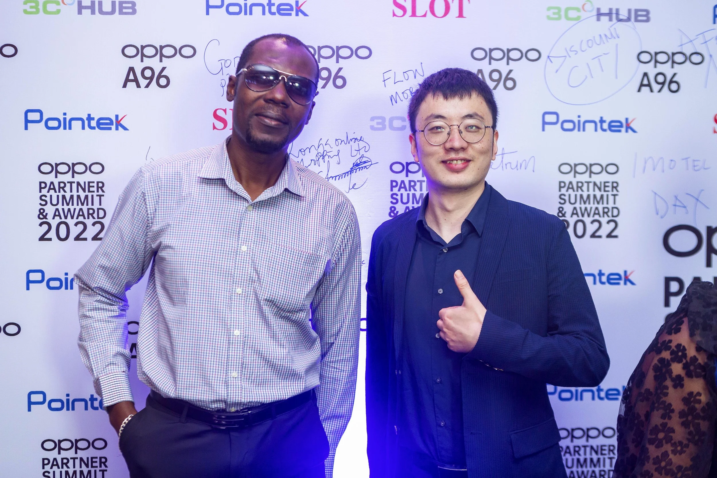 Two men standing together at a corporate event with a step-and-repeat backdrop featuring logos of Oppo, Pointek, 3C Hub, and SLOT. The man on the left is wearing sunglasses and a checkered shirt, while the man on the right is wearing glasses and a da