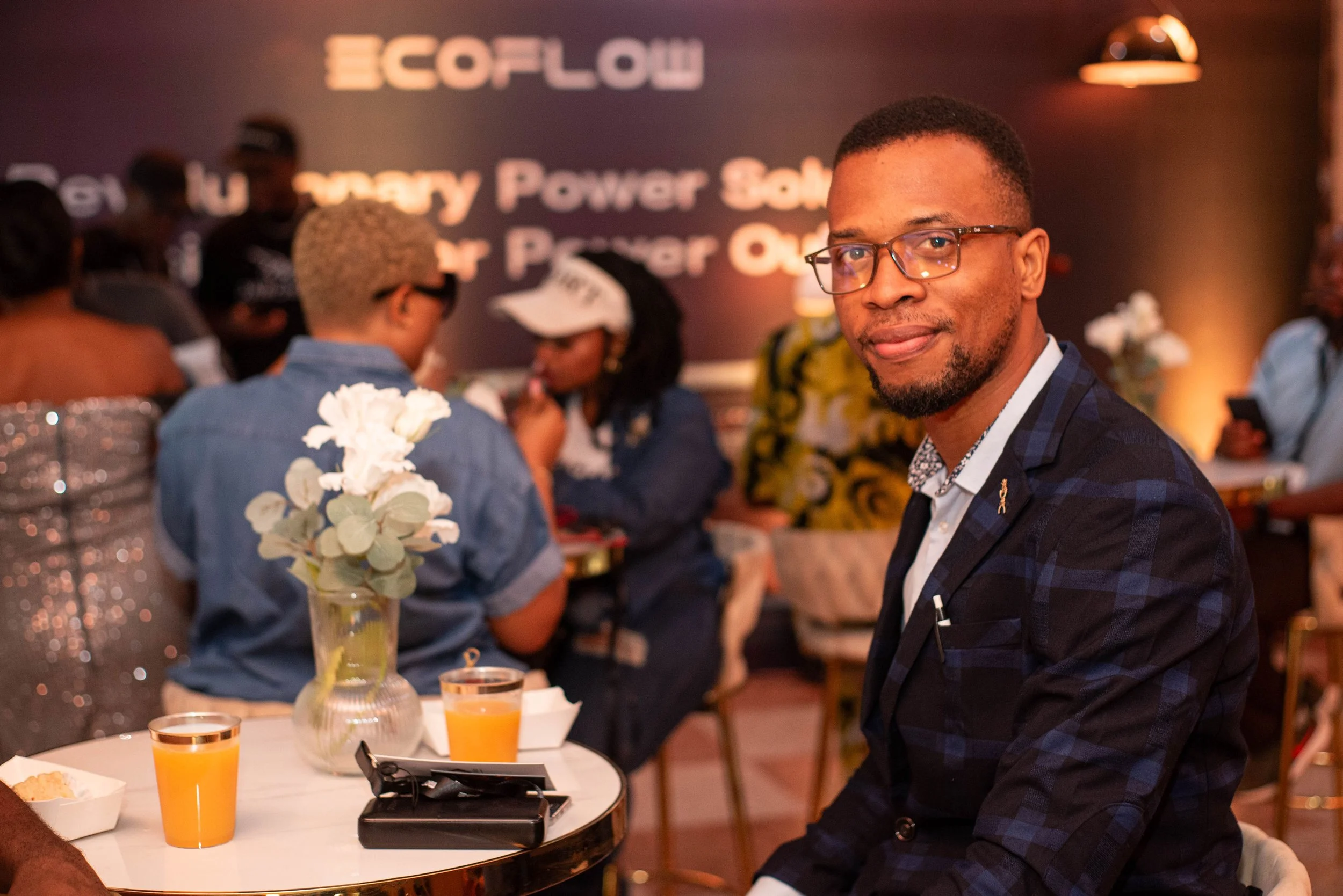 A man wearing glasses and a dark checkered suit smiling at the camera while sitting at a table. In the background, there are people gathered around, some wearing hats, and a wall with the text 'ECOFLOW' and a slogan about portable power solutions. Th