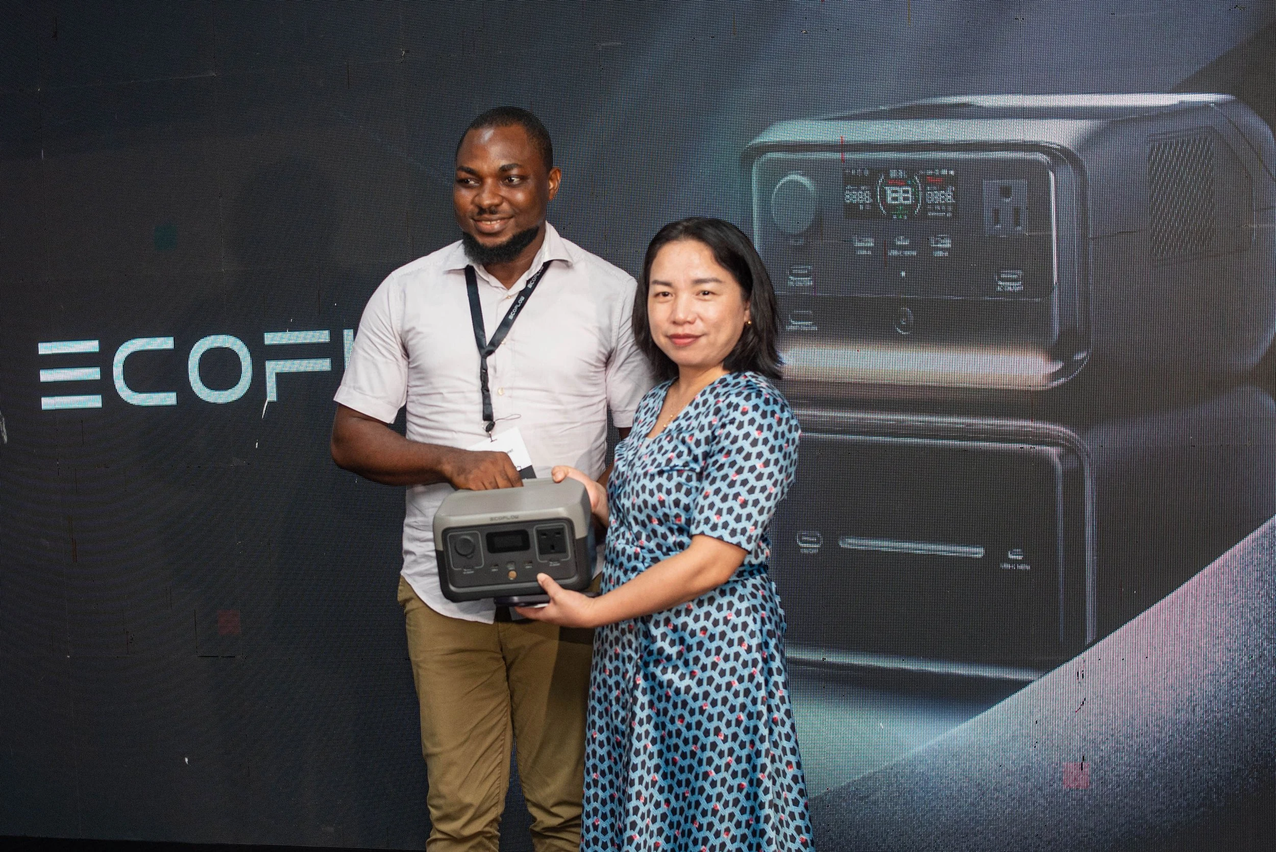 Two people shaking hands and exchanging a device in front of a digital screen with technical graphics.