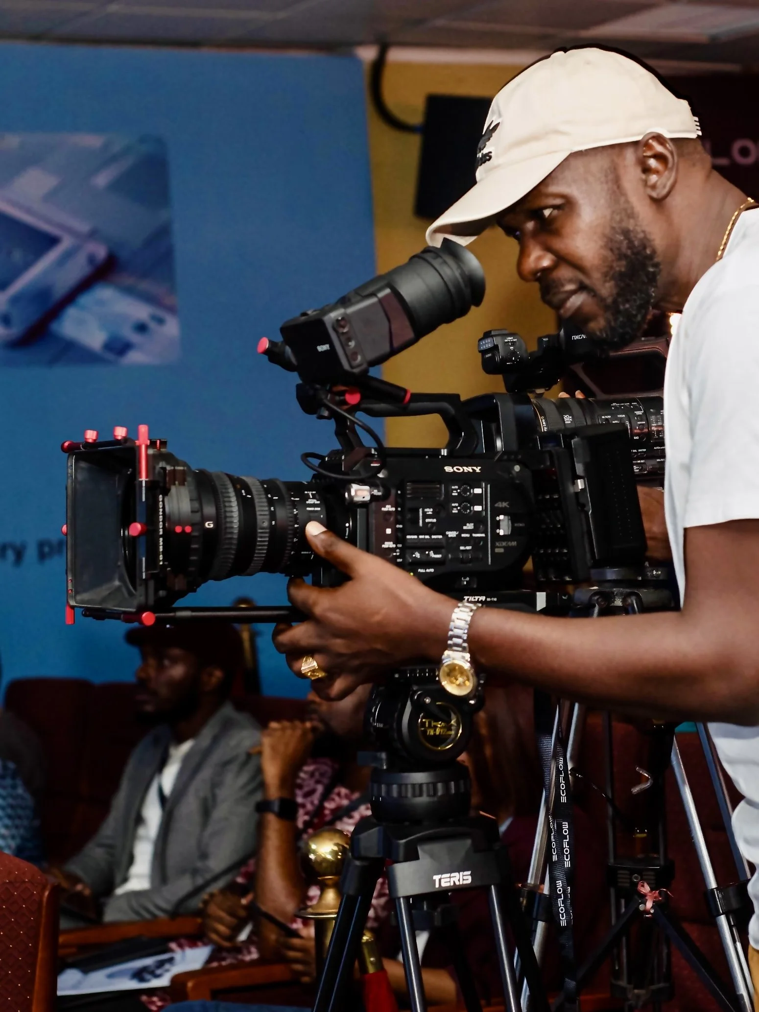 A man with a beard, white cap, and gold watch using a professional video camera on a tripod, with a woman sitting in the background.