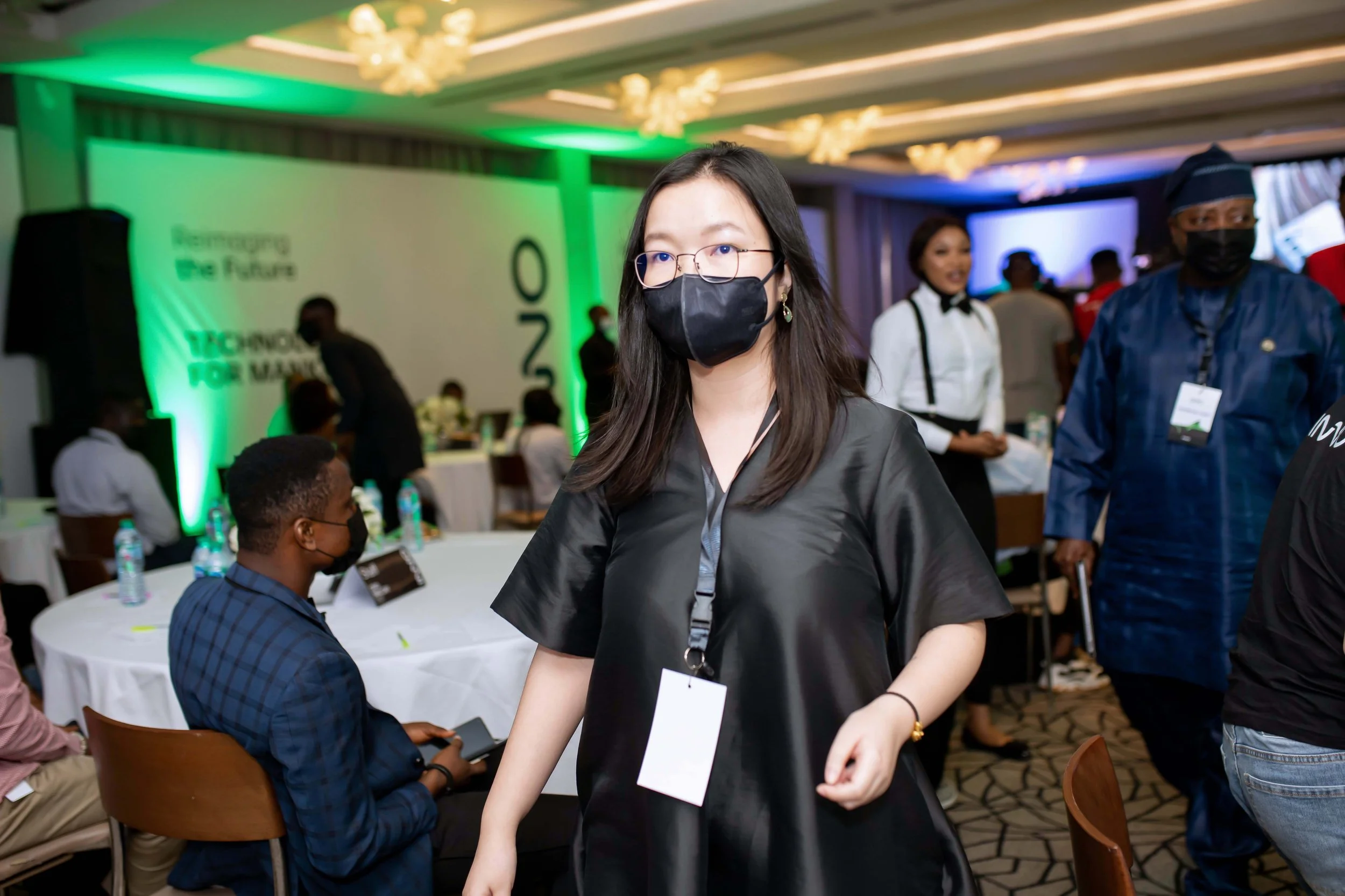 A woman in glasses and a black face mask walking in a conference room with other attendees, some wearing masks. The room has green and blue lighting, and a large display screen in the background.