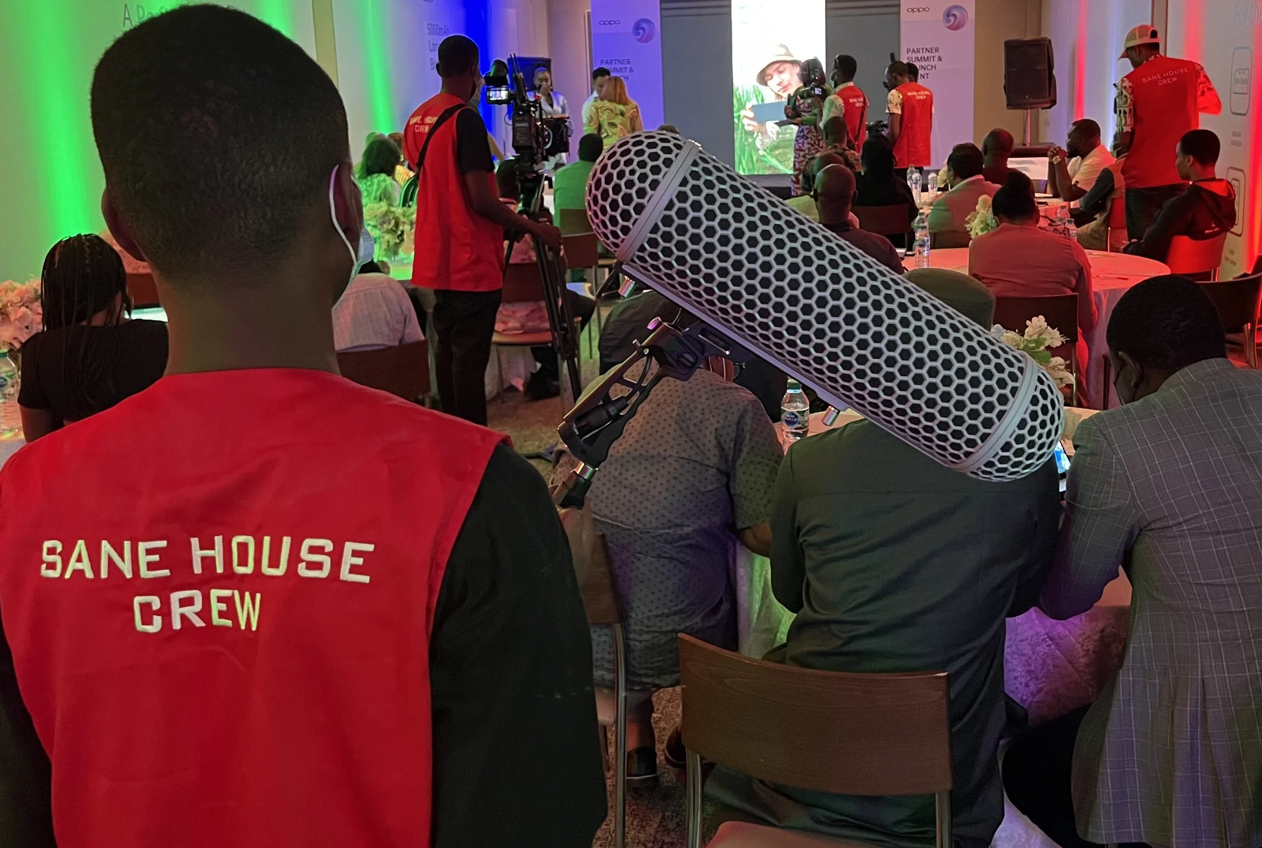 Photo of conference or seminar with attendees seated at tables, a stage with a screen and speakers, and security or event staff in red vests. The image includes a large microphone in the foreground, with colorful lighting and banners in the backgroun