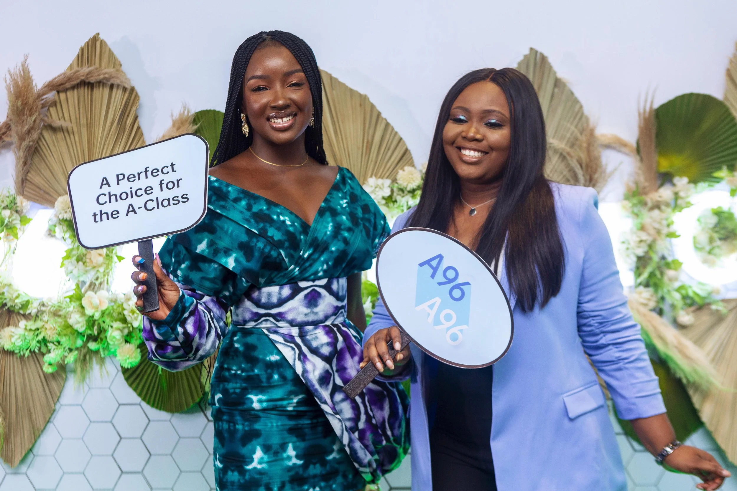 Two women standing together, holding signs, with a decorative backdrop of dried palm leaves and white flowers. One sign reads "A Perfect Choice for the A-Class." Both women are smiling, dressed stylishly, with one in a teal patterned dress and the ot
