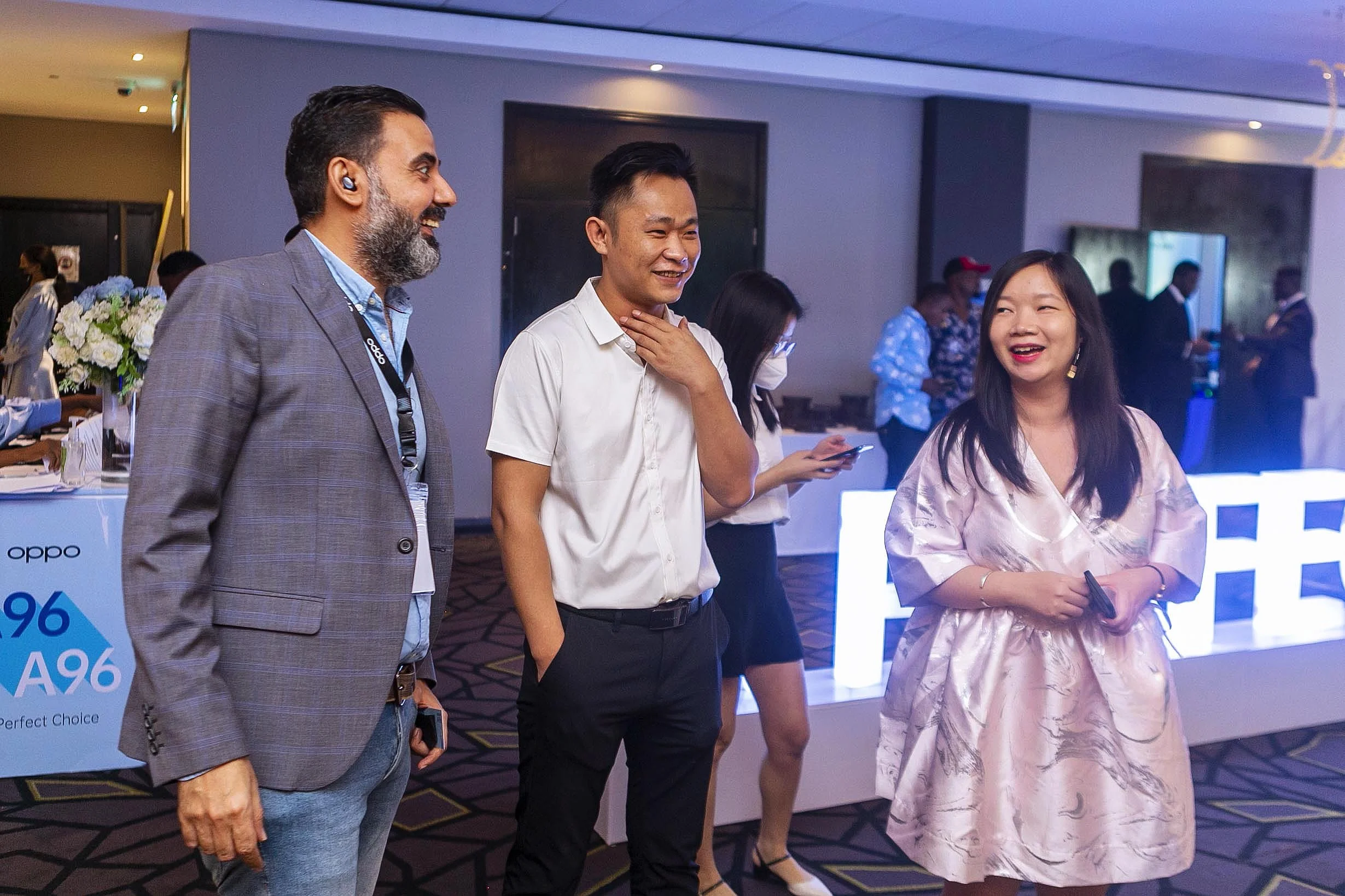Three people are standing and talking in a conference or event setting. The man on the left has a beard and gray hair, and is dressed in a gray blazer and blue shirt. The man in the middle, holding his hand to his throat, is smiling and wearing a whi