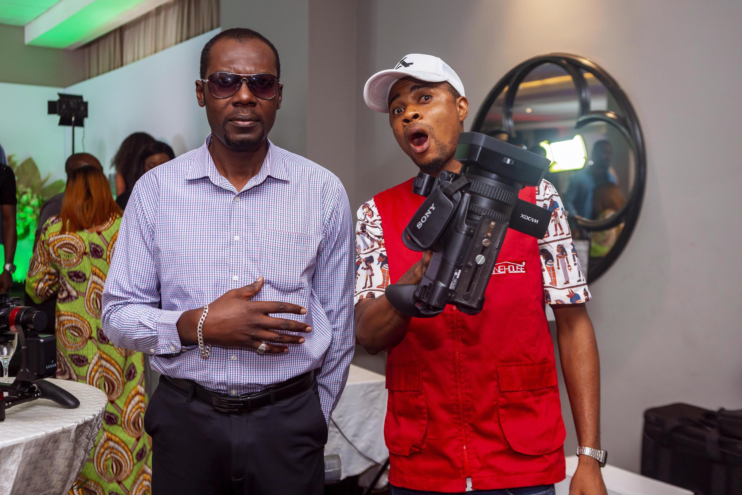 Two men at an indoor event, one with sunglasses and the other holding a camera, standing near a table with photography equipment. In the background, a woman with colorful clothing and other people are visible.