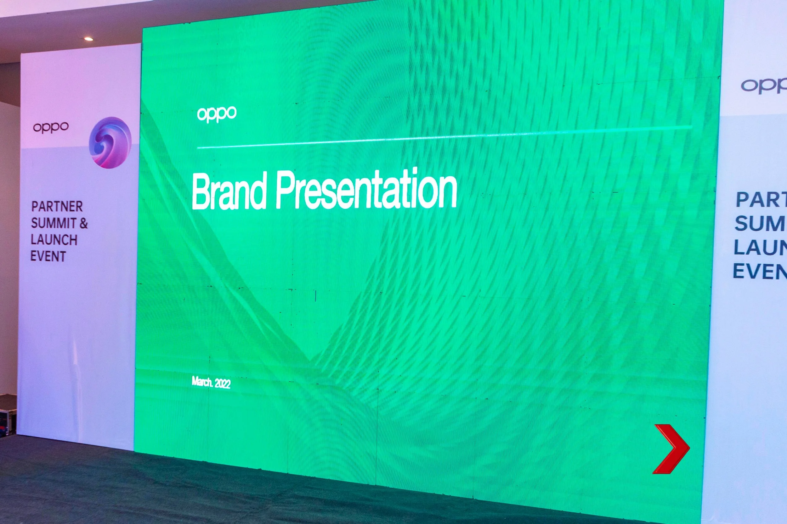 Large digital screen displays green background with white text that reads 'Brand Presentation'. On the left, there is a smaller partial screen with the Oppo logo and an abstract circular design. The bottom left corner of the screen displays the date March 2022. To the right, a red arrow points towards a slide or next part of the presentation. The setting appears to be a conference or event space.