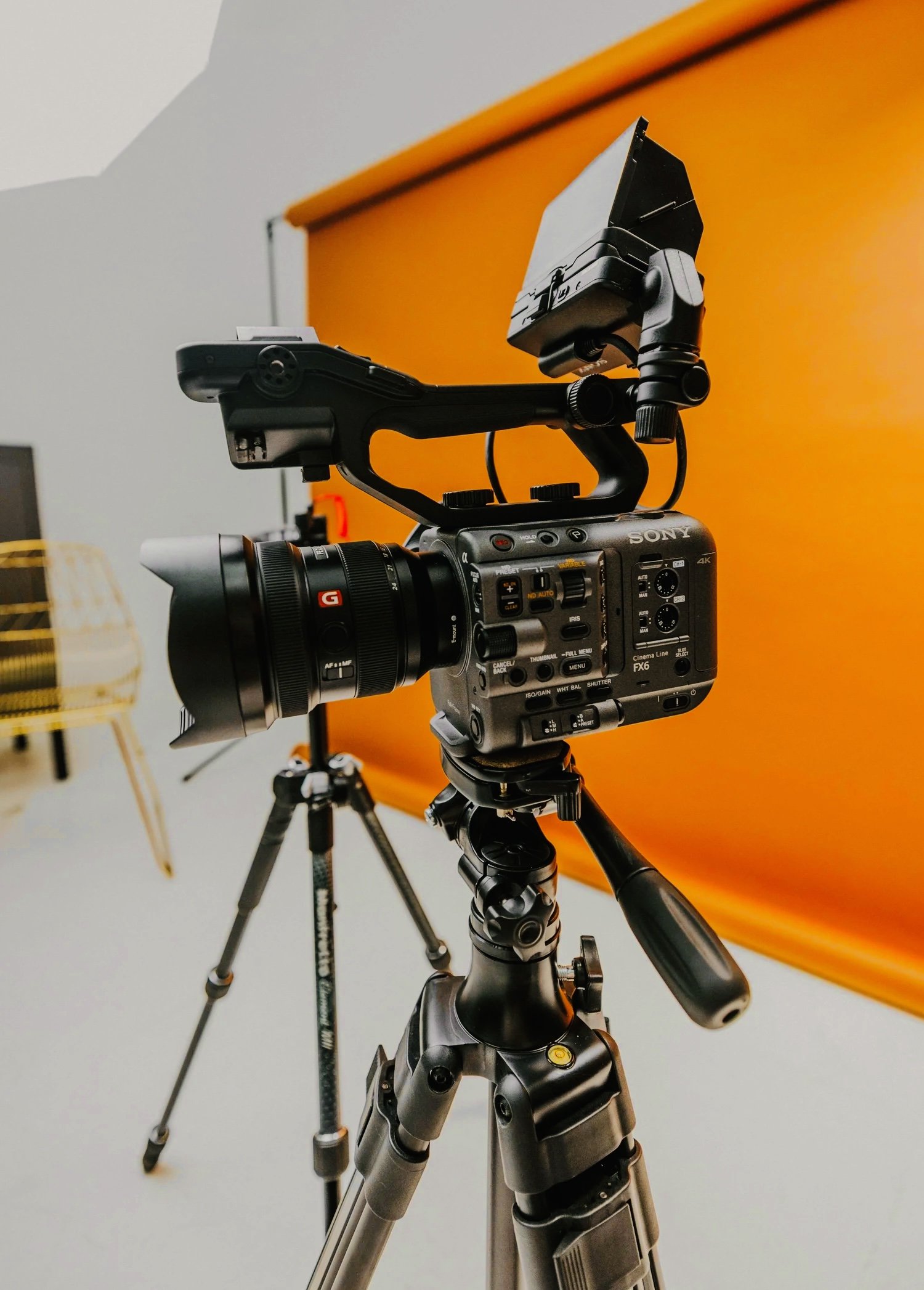 A professional video camera mounted on a tripod in front of an orange backdrop during filming or photography setup.