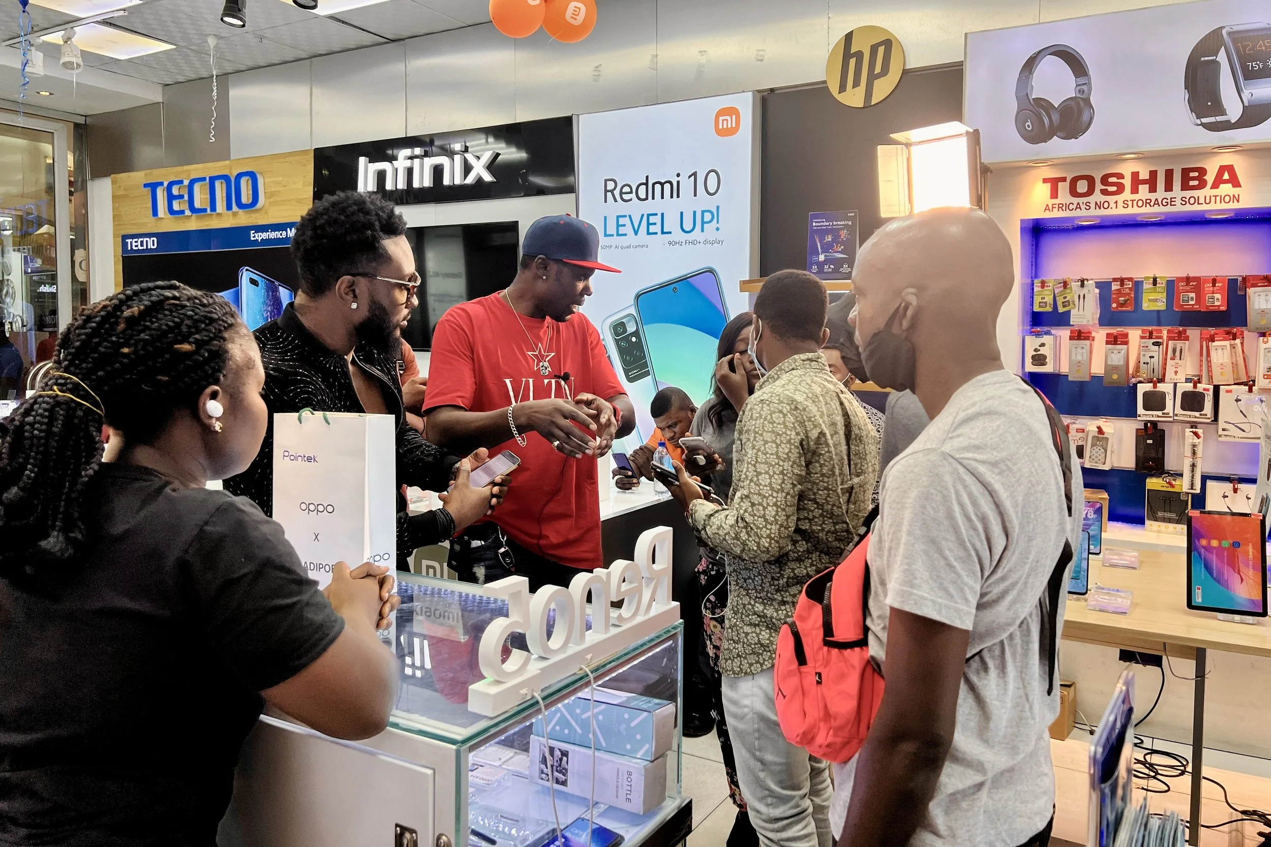 Sane House Media director speaking to cast and crew during a video shoot at a tech store, with phone and accessory displays in the background.