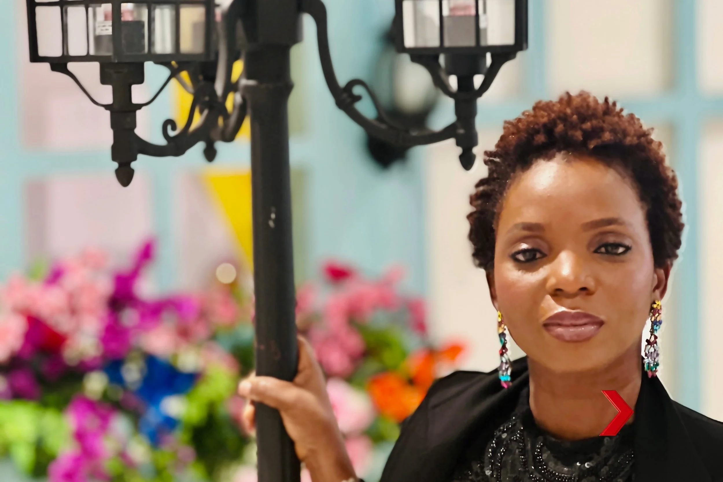 A woman with short curly hair, colorful earrings, and a black sequined top poses in front of a background filled with colorful flowers and a pastel-colored wall, holding onto a black structure.
