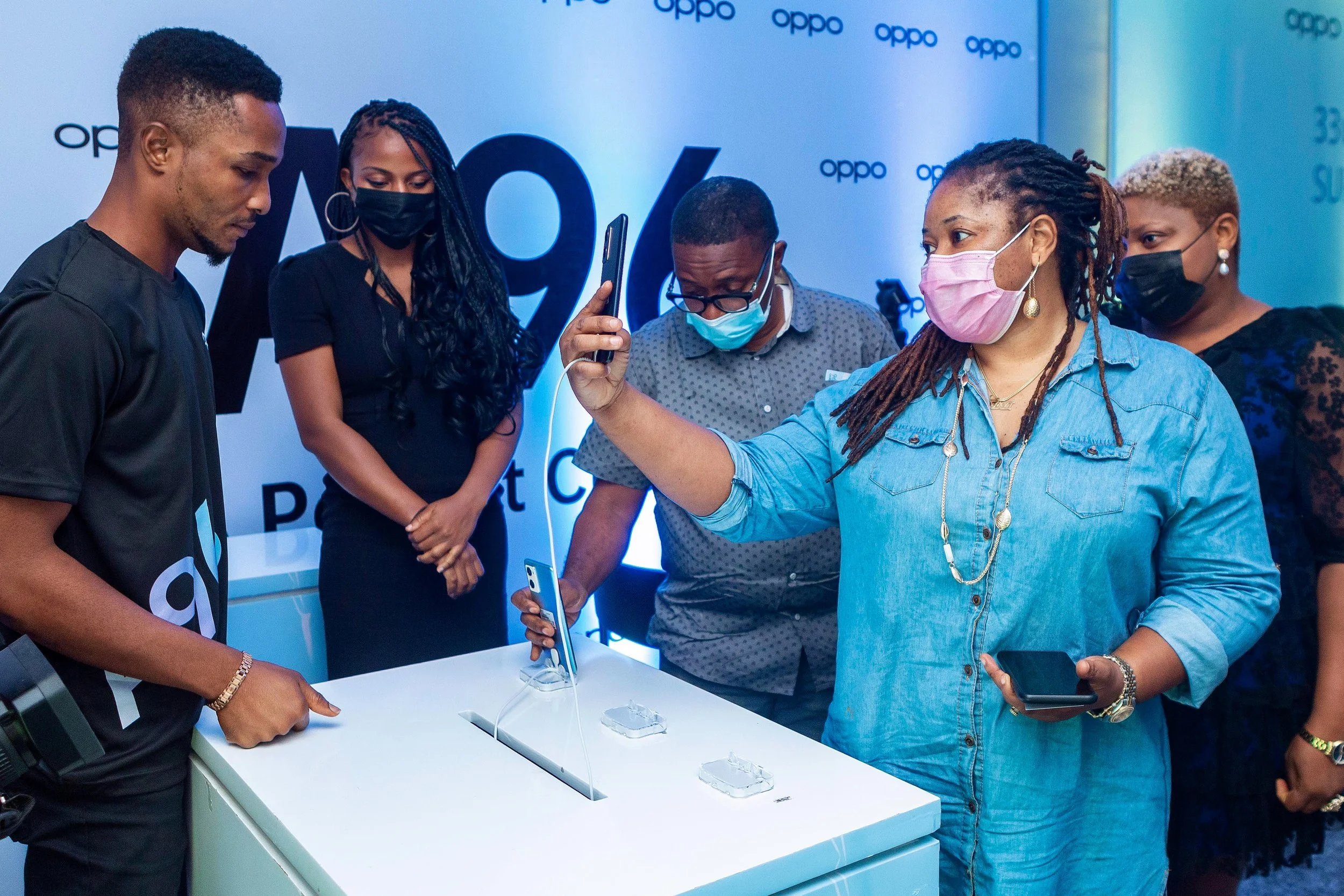 Group of people wearing face masks at an event, gathered around a display with smartphones, in front of a blue backdrop with 'oppo' logos.