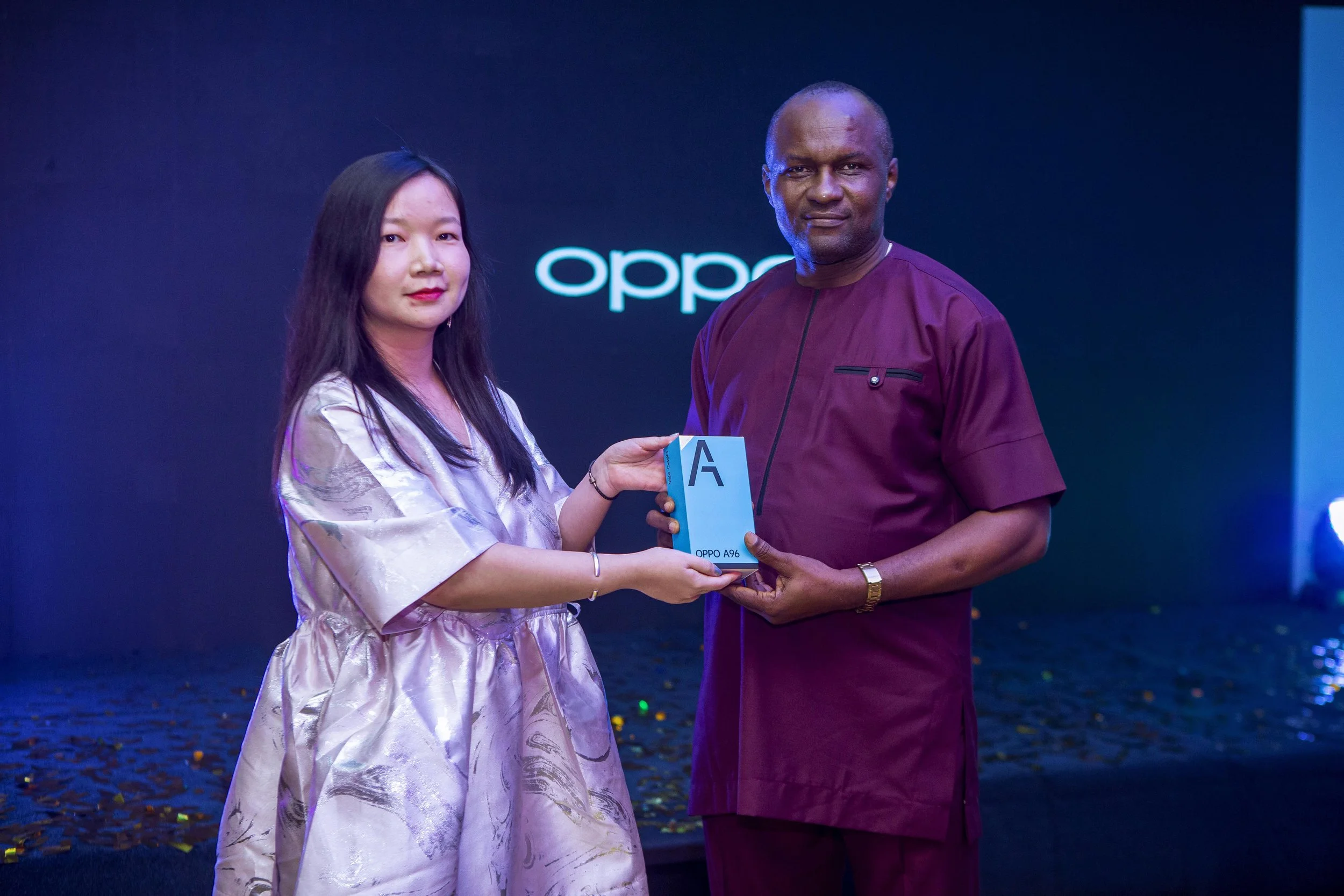 An OPPO executive and a guest standing together, with the woman handing the man a blue box labeled 'OPPO A96', during an event with a dark background and the word 'oppo' illuminated in blue.
