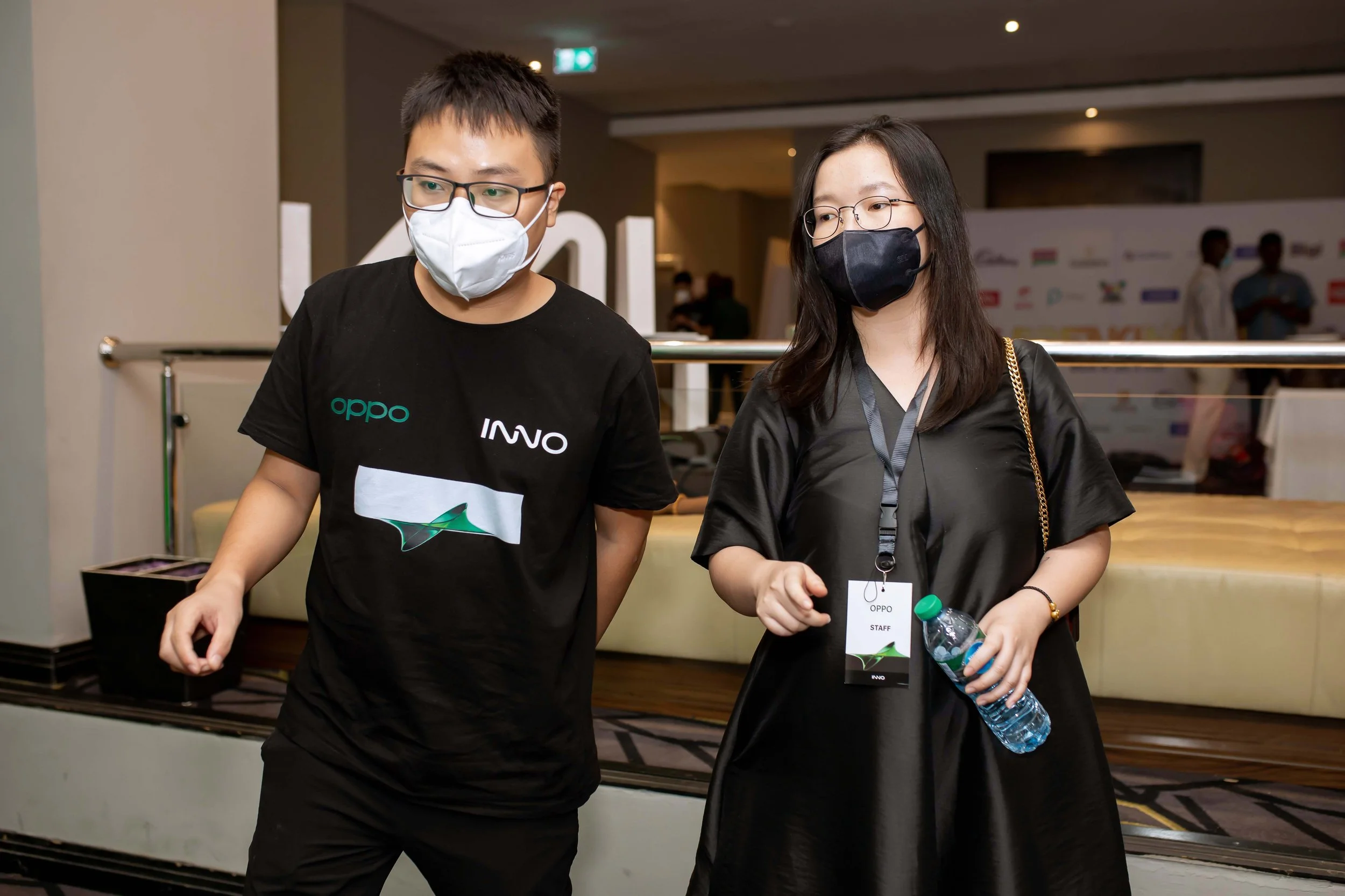 Two OPPO staff wearing face masks walking indoors at an OPPO event, with a background showing a wall with logos and other attendees.
