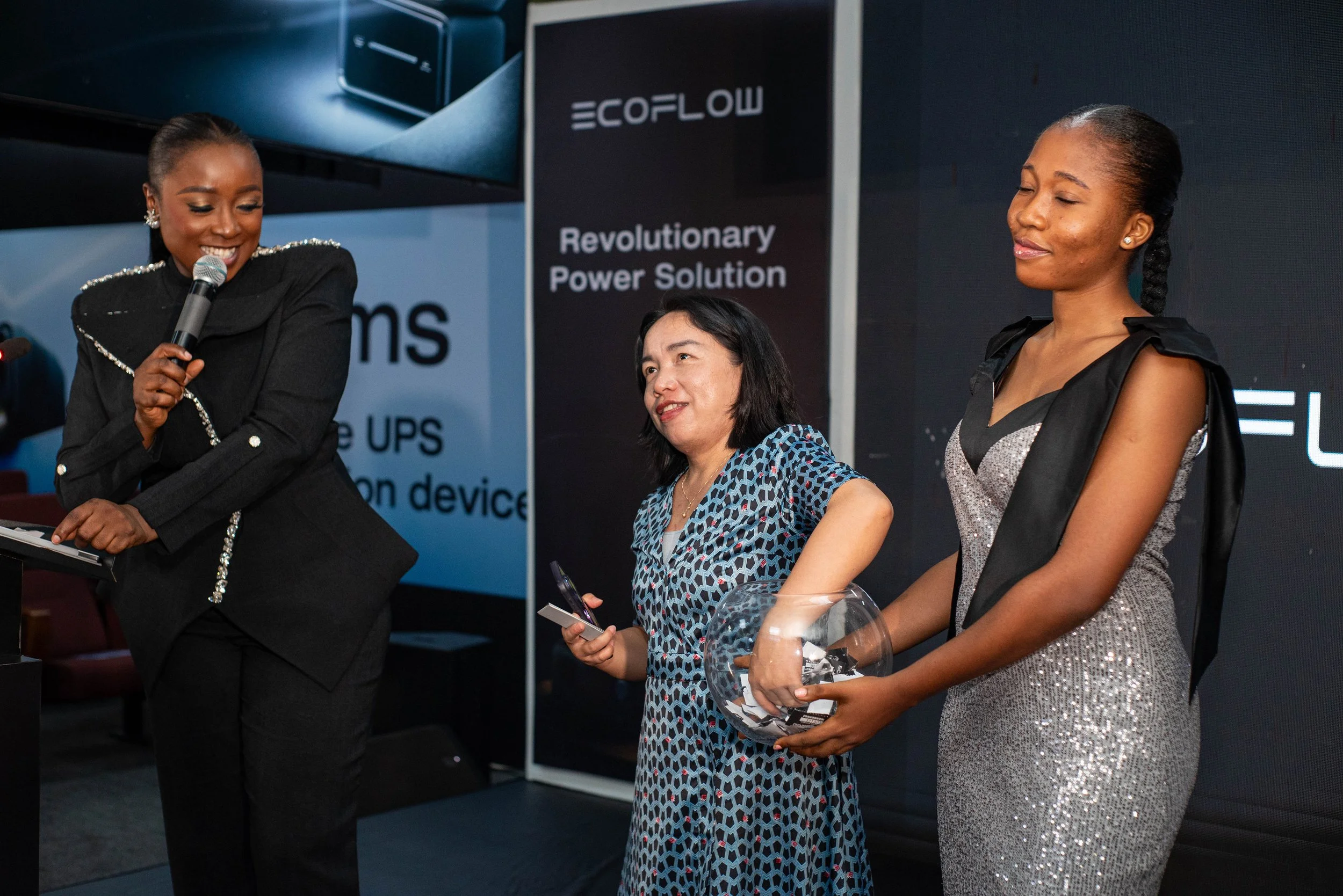 Three women standing and engaging in conversation at a professional event, with a large monitor and signage in the background.