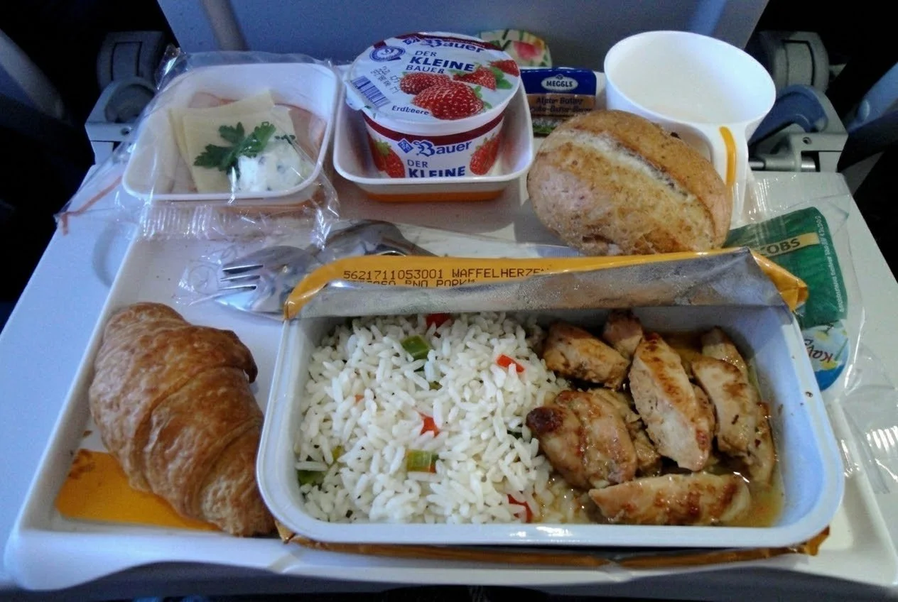 An overhead photograph captures a traditional economy class in-flight meal tray on an airplane, featuring a peeled-back hot dish revealing chicken and rice, accompanied by a strawberry yogurt, a bread roll, a croissant, cheese, and butter.