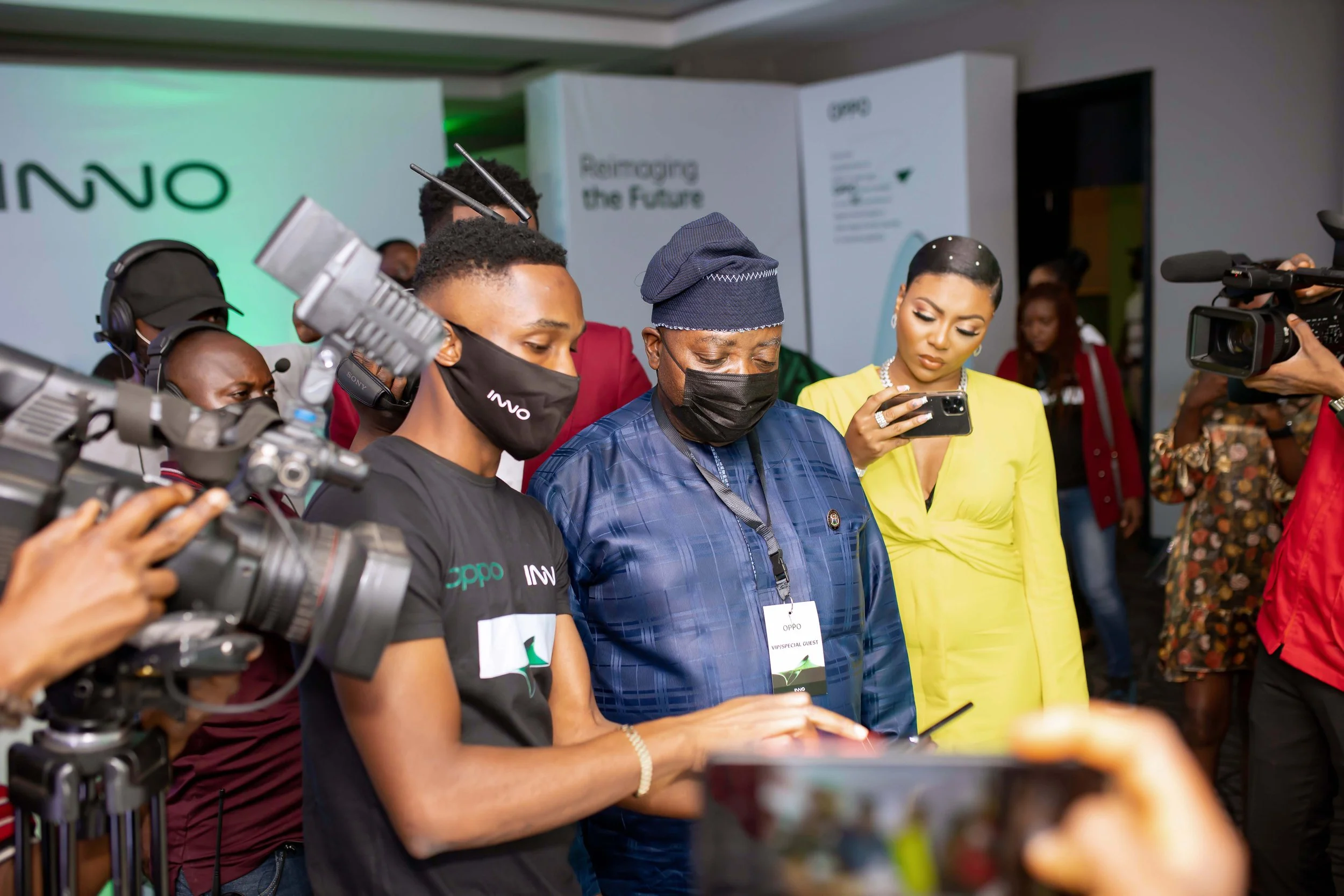 A group of people at an event, some wearing face masks, engaging with electronic devices and a camera. There is a background with the words "Reimagine the Future" .