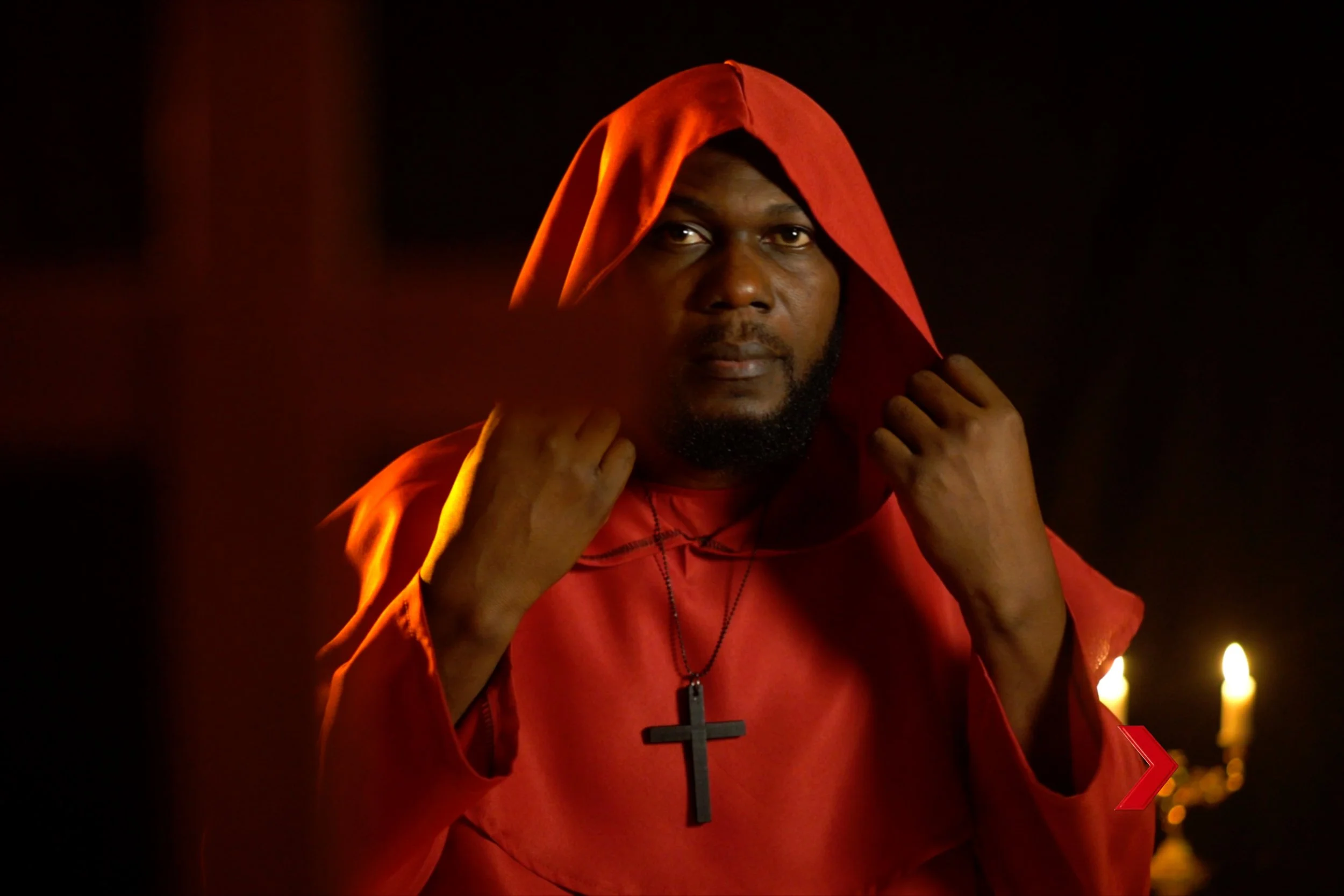 Rap artist Nutty Pin wearing a red hoodie and black cross necklace, holding the hoodie with both hands. The background is dark with some lighted candles.