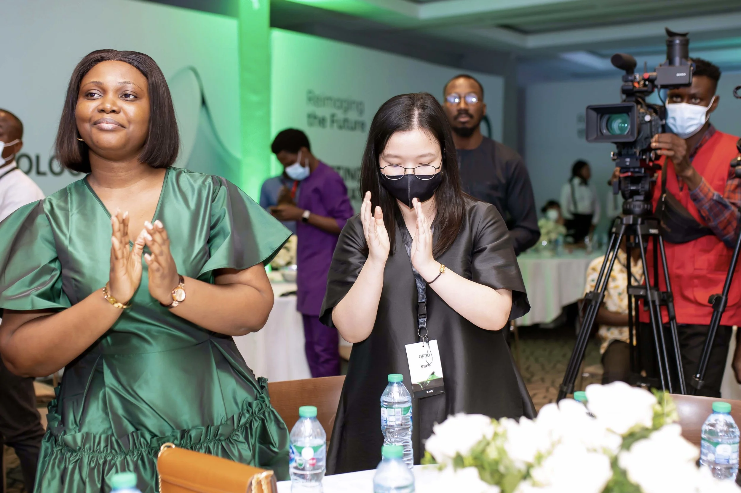 People attending an event, with some wearing masks. A woman in a green dress and a woman in a black shirt are in the foreground, with the woman in black showing a praying gesture. A camera person is filming in the background.