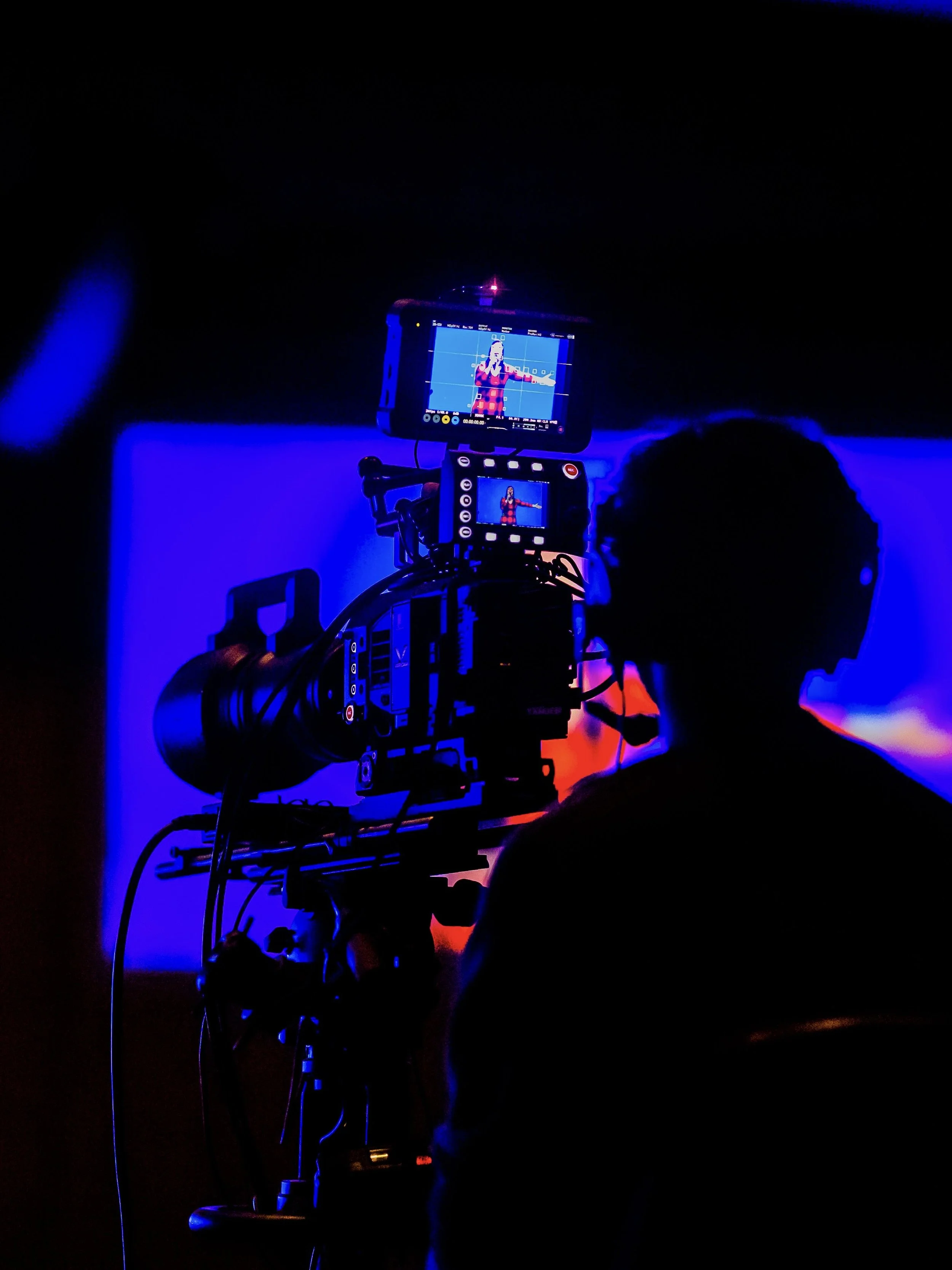 A person operating a camera on a tripod capturing a woman on stage in a dark setting with blue lighting.
