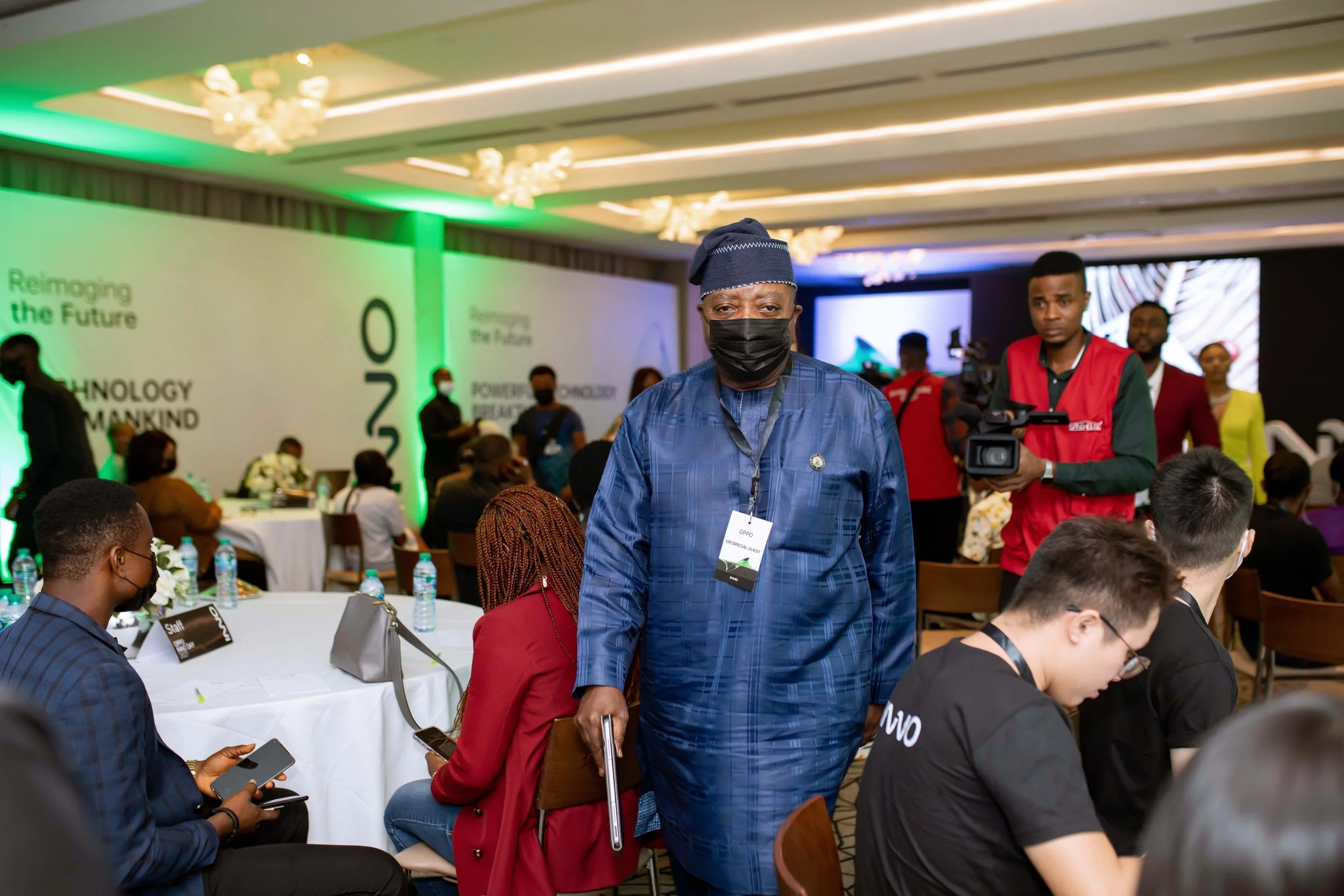 A man wearing a face mask, traditional clothing, and an event badge stands amidst a conference or seminar. The room is filled with people seated at tables, some engaging with electronic devices. The background features a large screen and a wall with 