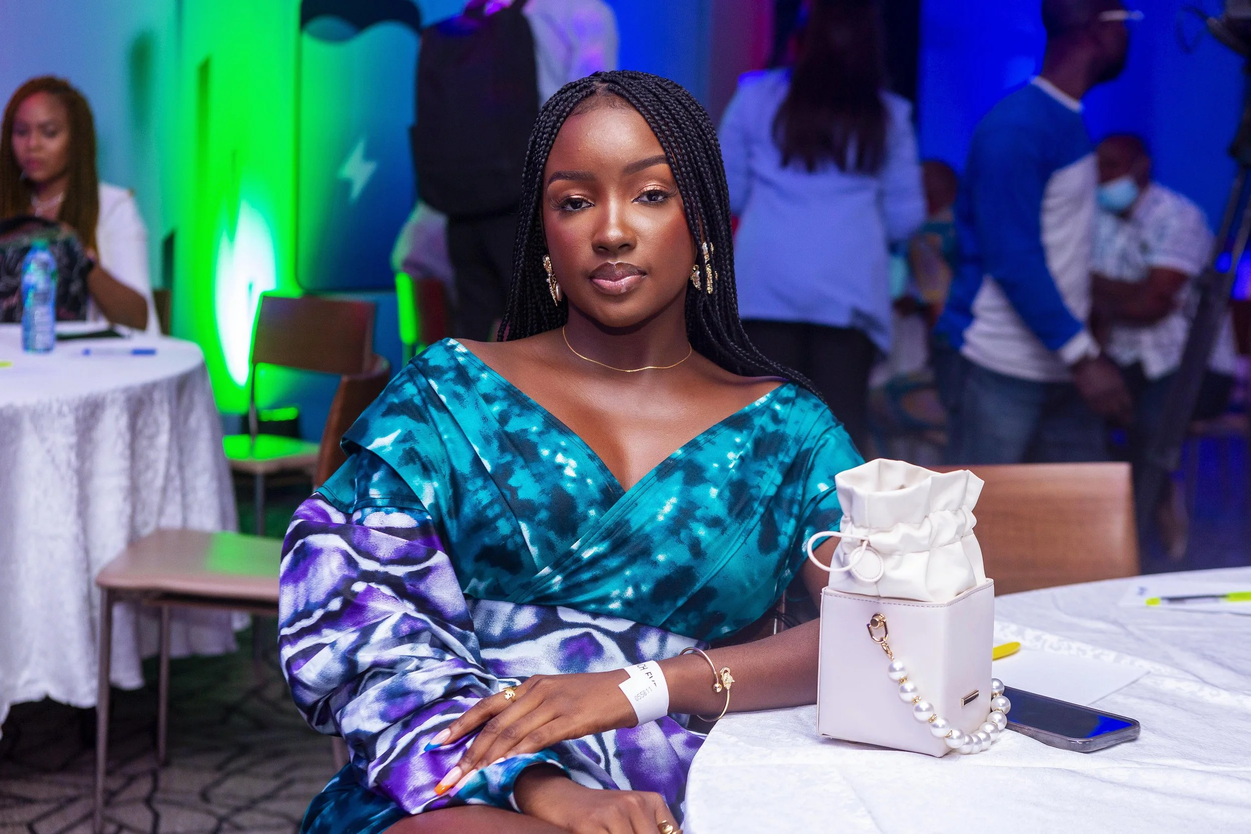 A young woman with braided hair and earrings sitting at a table in a colorful event space with other people in the background. She is wearing a blue patterned dress and has a white purse, a smartphone, and a wristband on her arm.