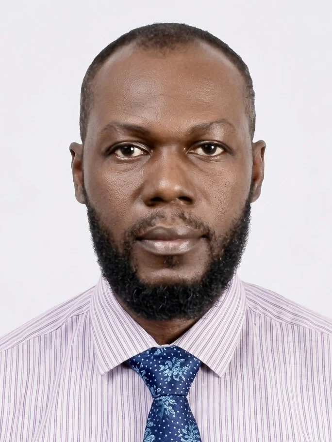 Portrait of a man with dark skin, short hair, and a beard, wearing a light purple striped dress shirt and a blue patterned tie, against a plain white background.
