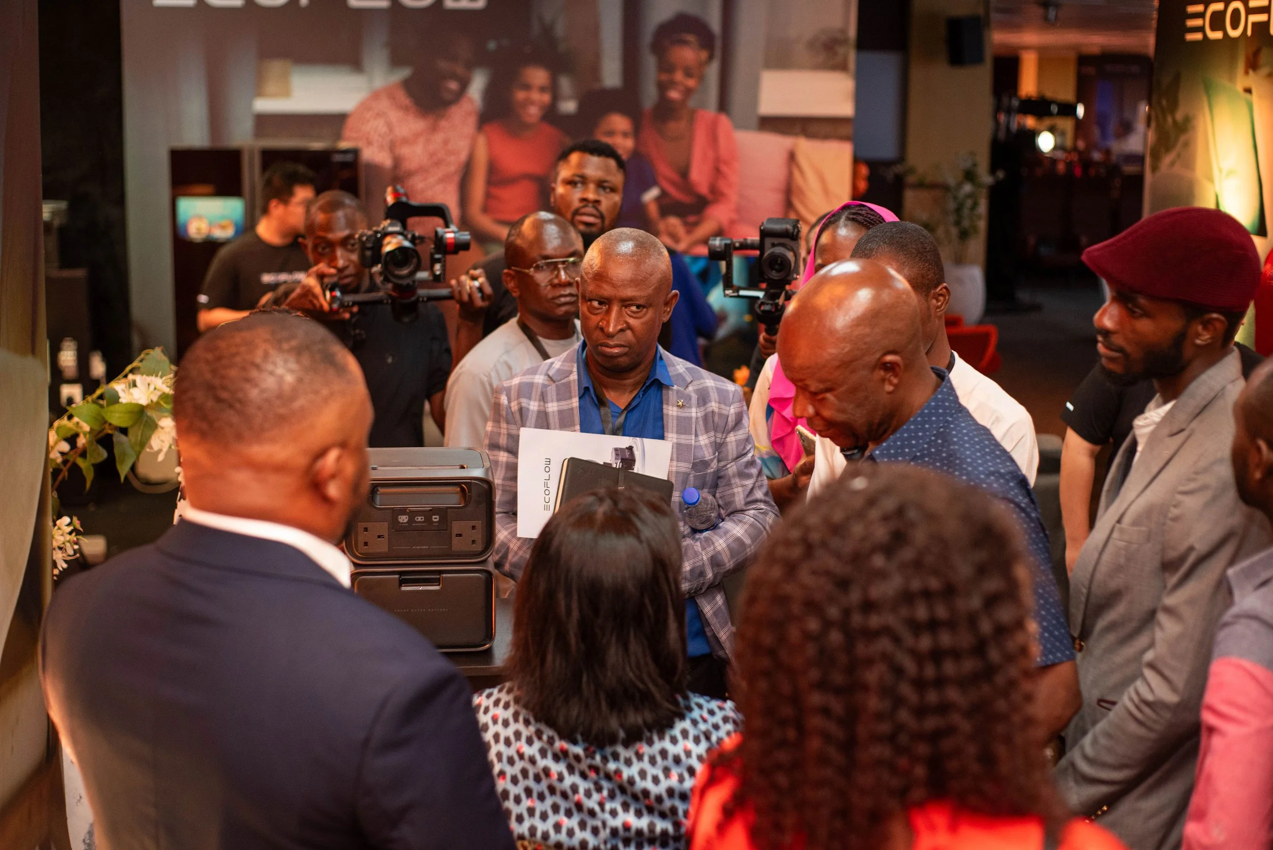 Guests gathered around a table, with some holding cameras, listening to a person speaking at tan EcoFlow event. In the background, a large poster with smiling faces and the word 'ECO' is visible.
