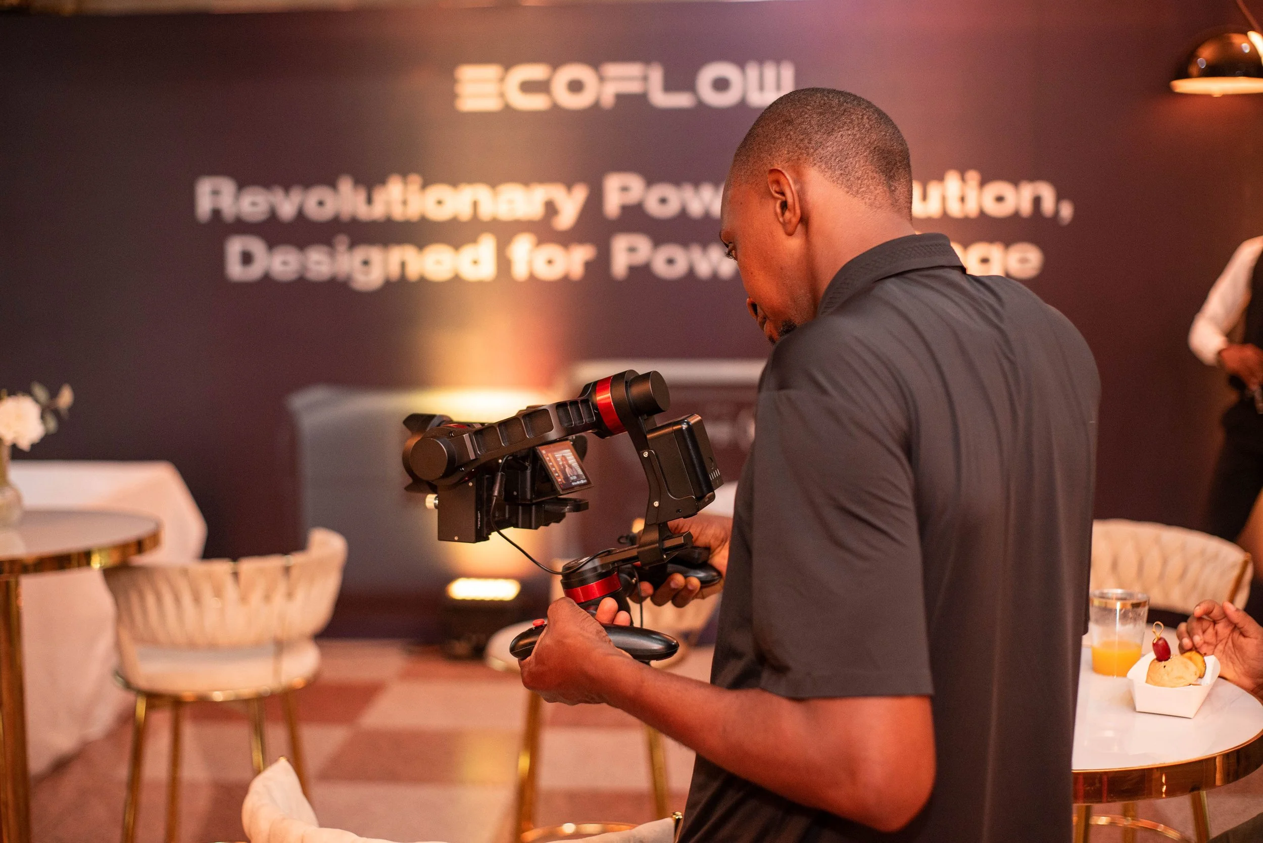 A man setting up a camera on a tripod at an indoor event with a sign reading 'ECOFLOW' and a tagline. There are tables with food and drink on the side.