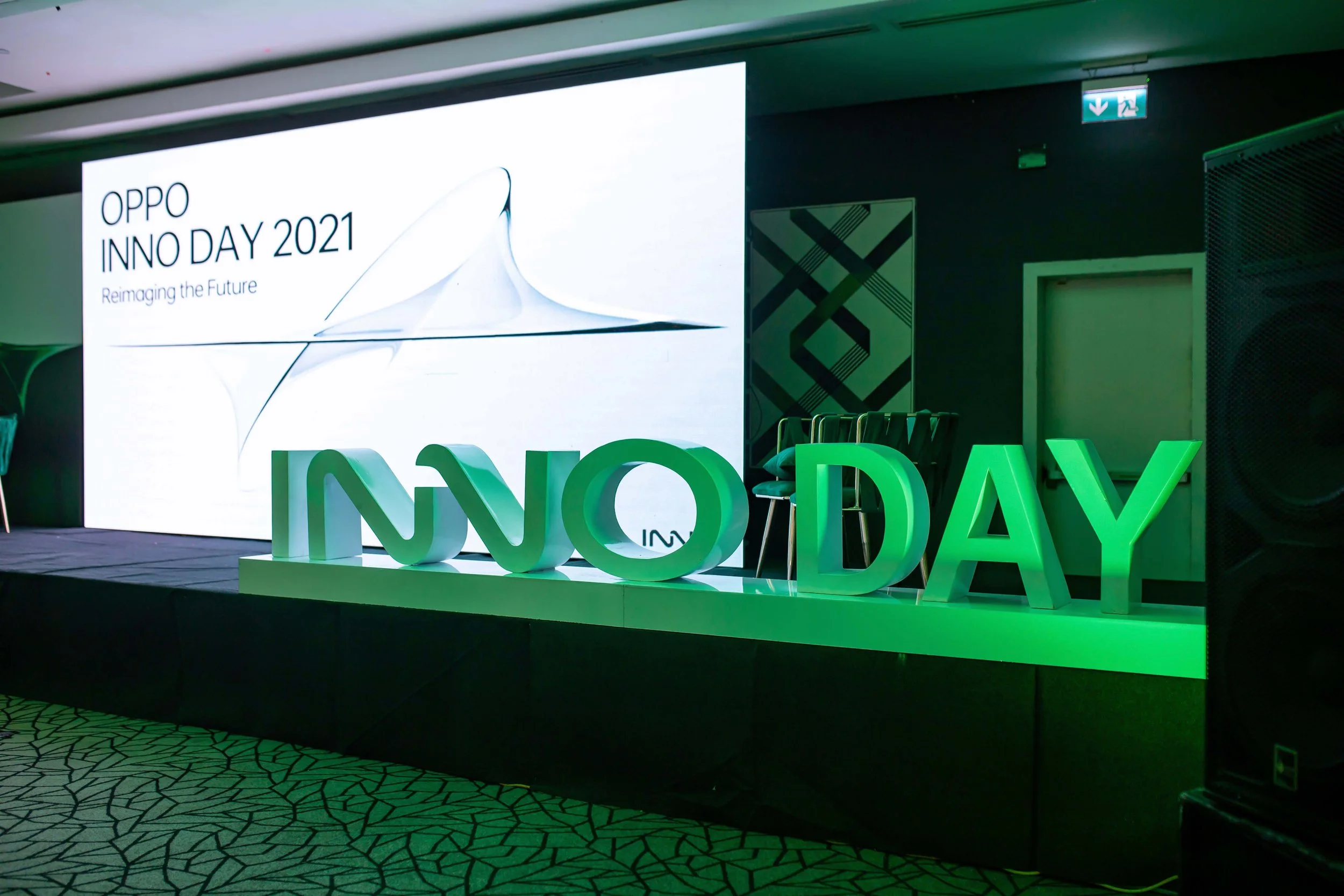 A stage at OPPO INNO DAY 2021 with a large screen displaying the event name, and large green illuminated letters spelling 'INNO DAY' on the stage. Chairs are lined up on the stage, and there is a patterned carpet on the floor.