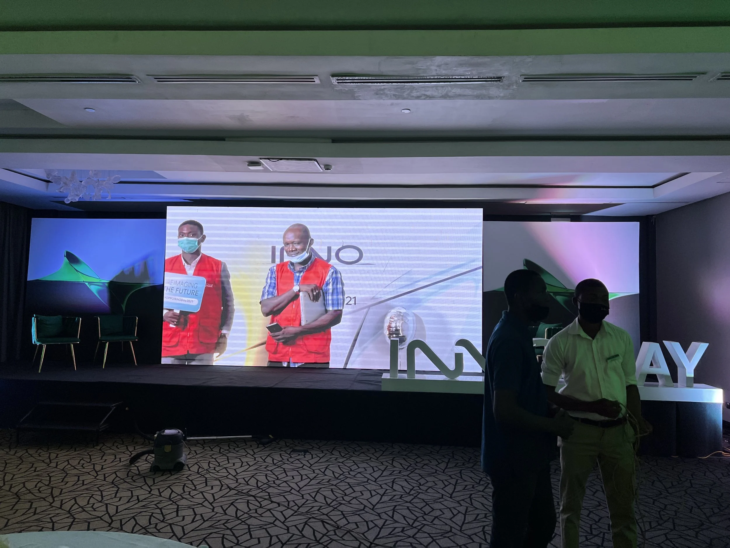 Sane House Media crew members wearing red vests and face masks, on stage screen at an event. 