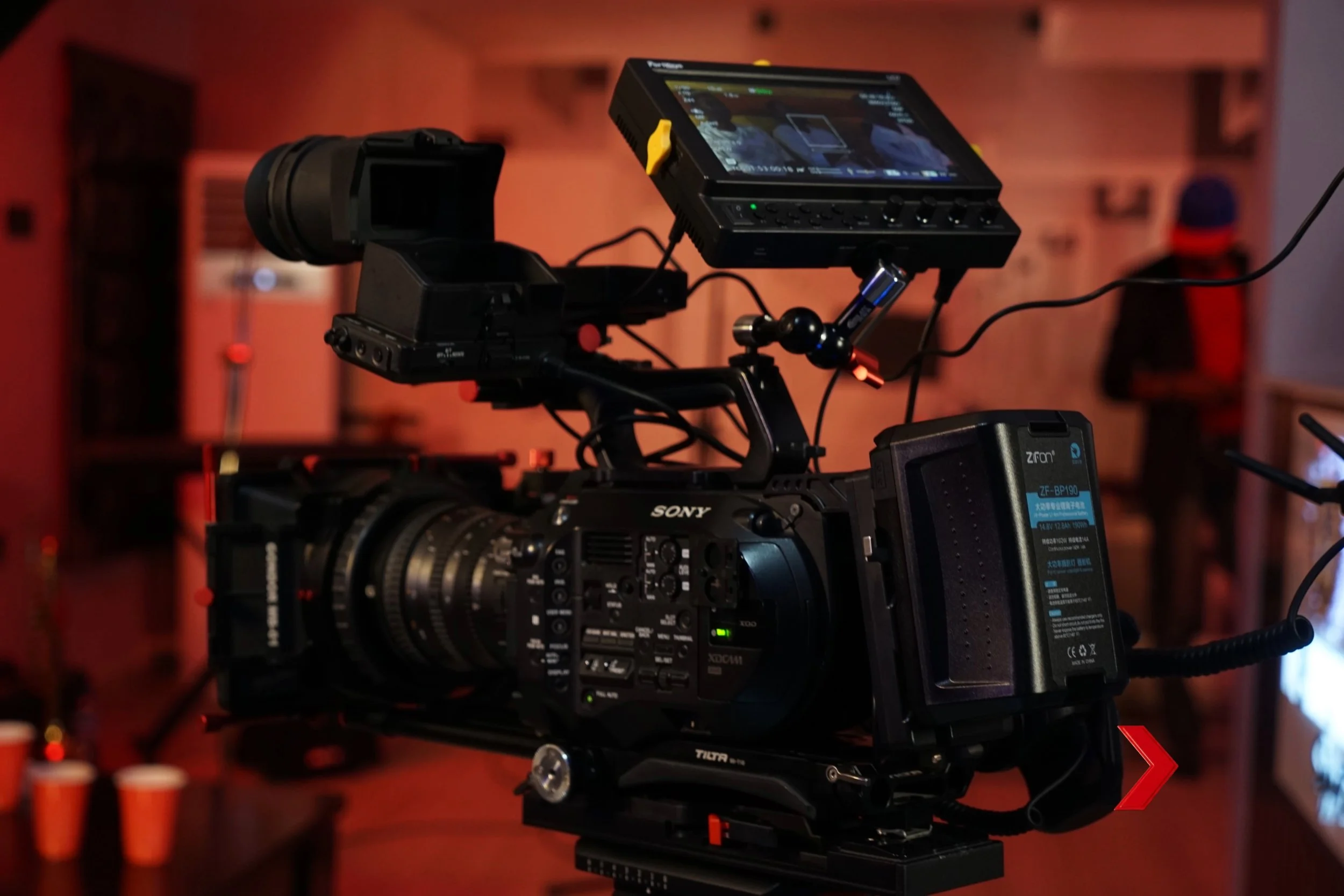 Close-up of aSony FS7 professional video camera on a tripod with a monitor attached, in a dimly lit indoor setting with red lighting.