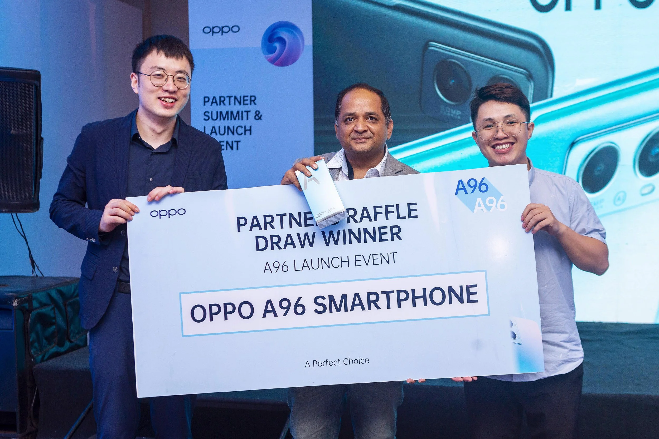 Three men holding a large check that reads "Partner Raffle Draw Winner" and "OPPO A96 Smartphone" at an event. The background features a poster with the OPPO logo and a large image of a smartphone.