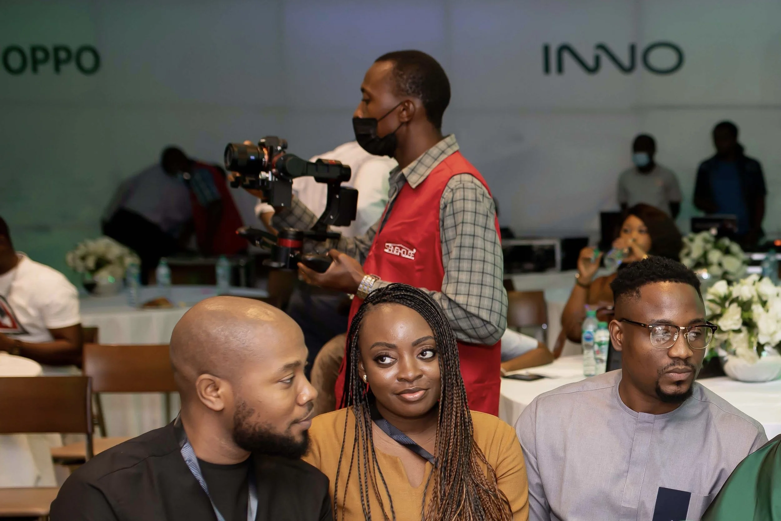 A person standing with a camera on a gimbal, wearing a red vest and face mask, at a conference or event with people seated around and a background with the "OPPO" and "INNO" logos.