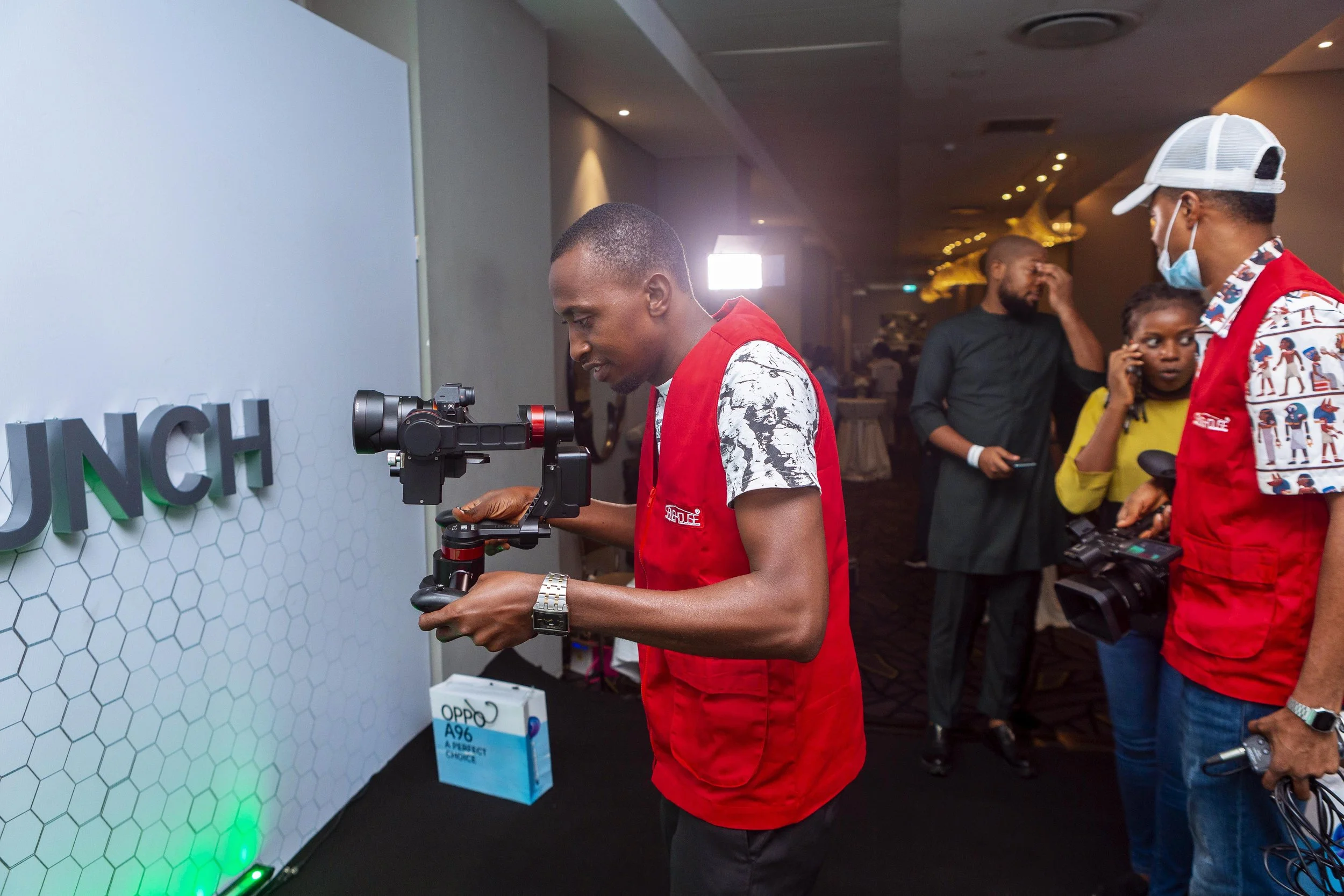 A man in a patterned shirt and red vest is operating a camera mounted on a gimbal at an event. Other attendees, including a woman on the phone and another man in a red vest with a patterned shirt and face mask, are visible in the background. There is