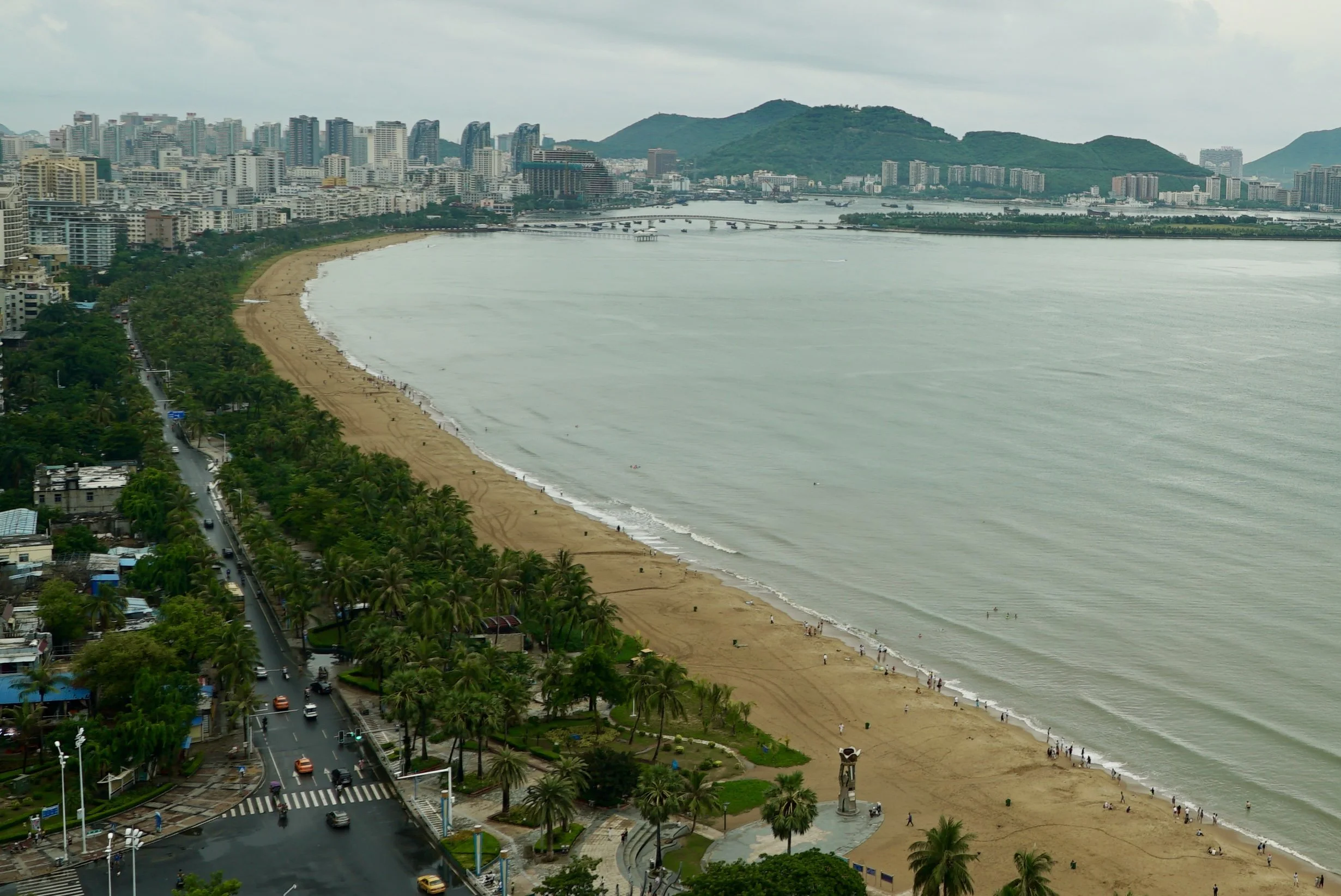 A High-angle view of Sanya Bay beach, a palm-lined road with cars, and distant city buildings.