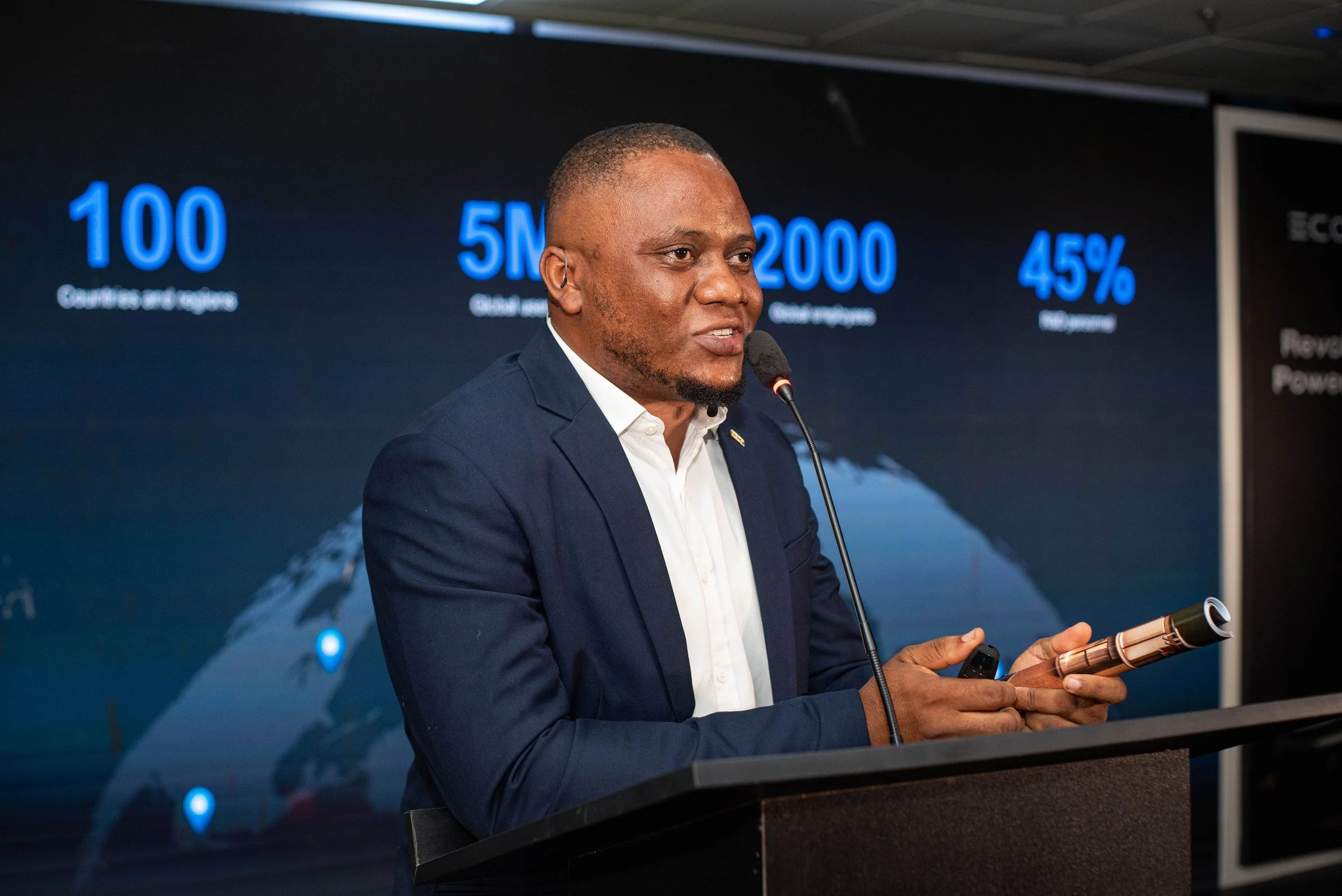 A man in a navy blazer and white shirt standing at a podium, speaking into a microphone, with a large presentation screen behind him displaying statistics related to EcoFlow data.