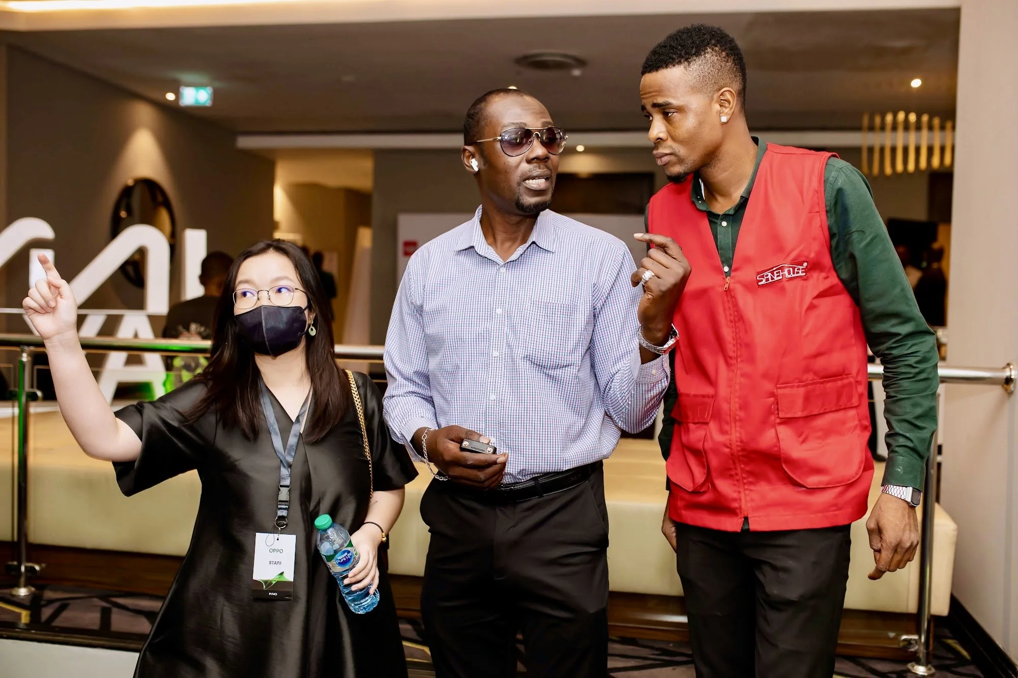 Three people engaged in conversation at an indoor event. The woman on the left, wearing glasses, a black mask, and a black dress, holds a water bottle and has an OPPO staff badge. The man in the middle, wearing glasses and a checkered shirt, holds a 