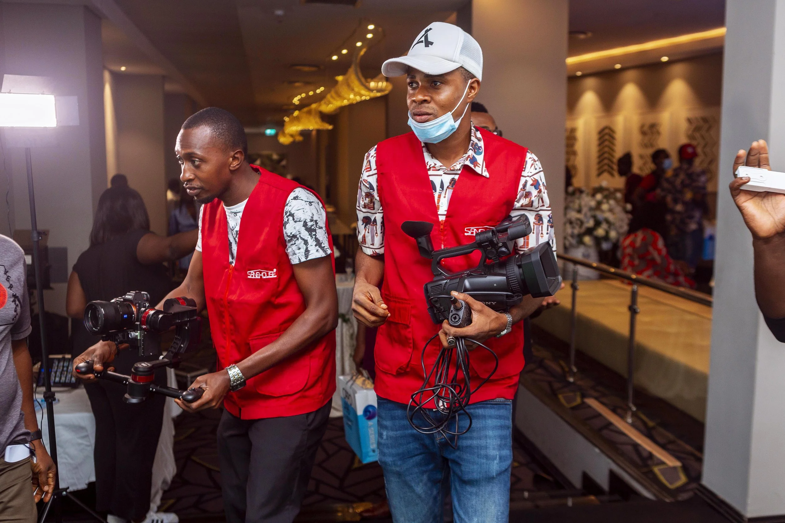 Two men in red vests are operating camera equipment at an indoor event, with one wearing a white cap and a face mask hanging below his chin, and the other looking at a camera in his hands.