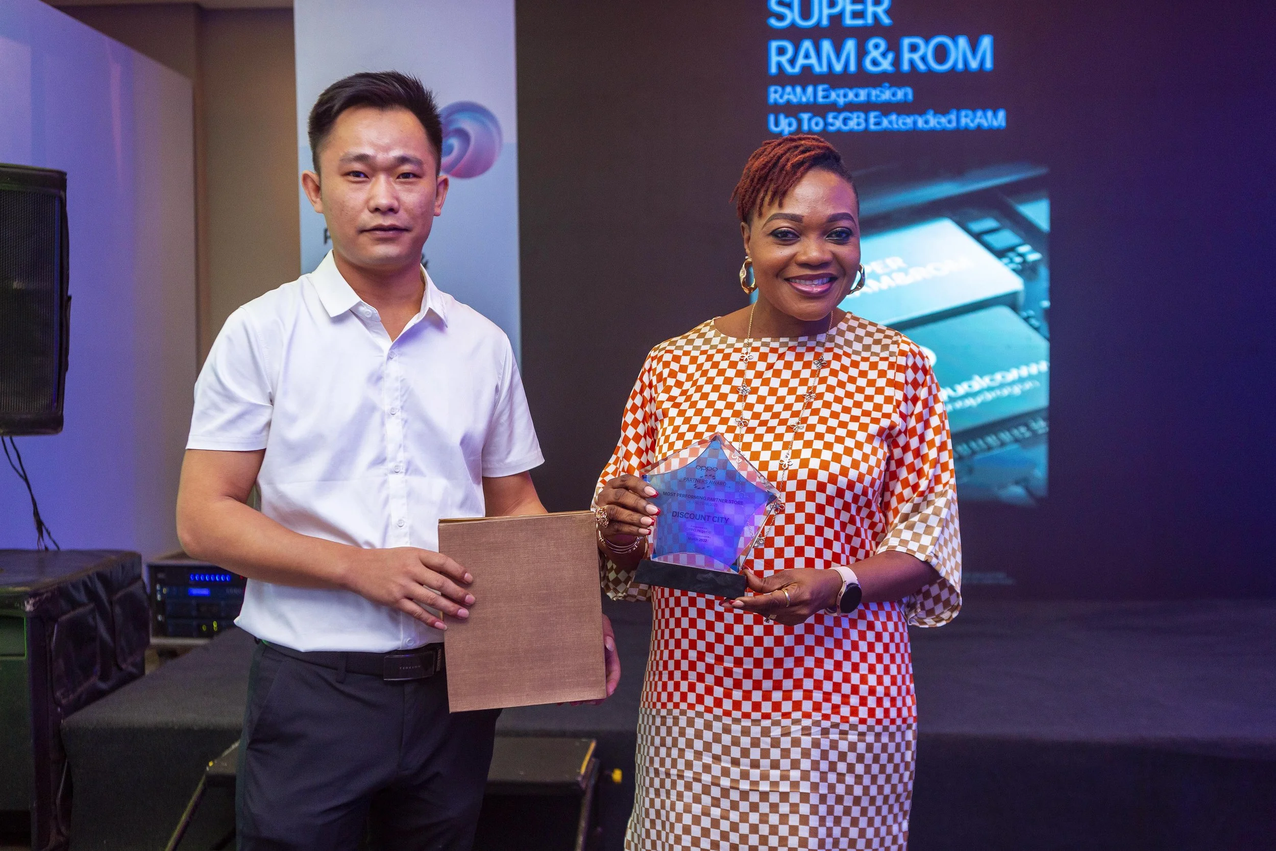 Two people standing on a stage at an award event, holding awards. The man on the left has short dark hair, wearing a white shirt, and the woman on the right has short, reddish-brown hair, wearing earrings, a checkered dress, and a necklace. There is 