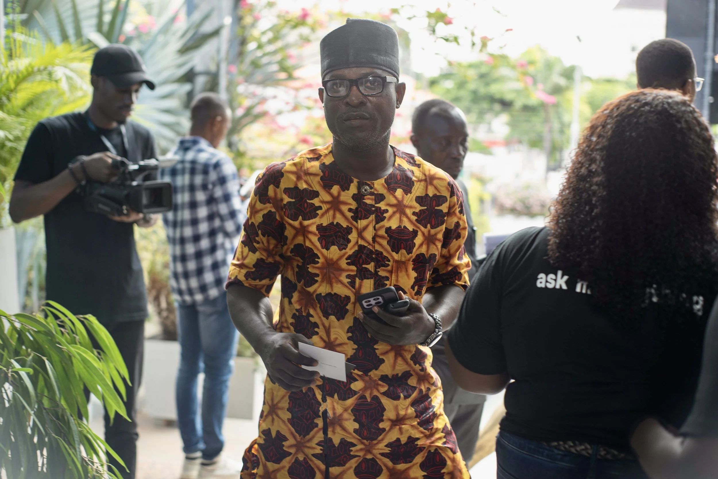 A man wearing glasses and a traditional African shirt holding a phone, standing outdoors with other people in the background.