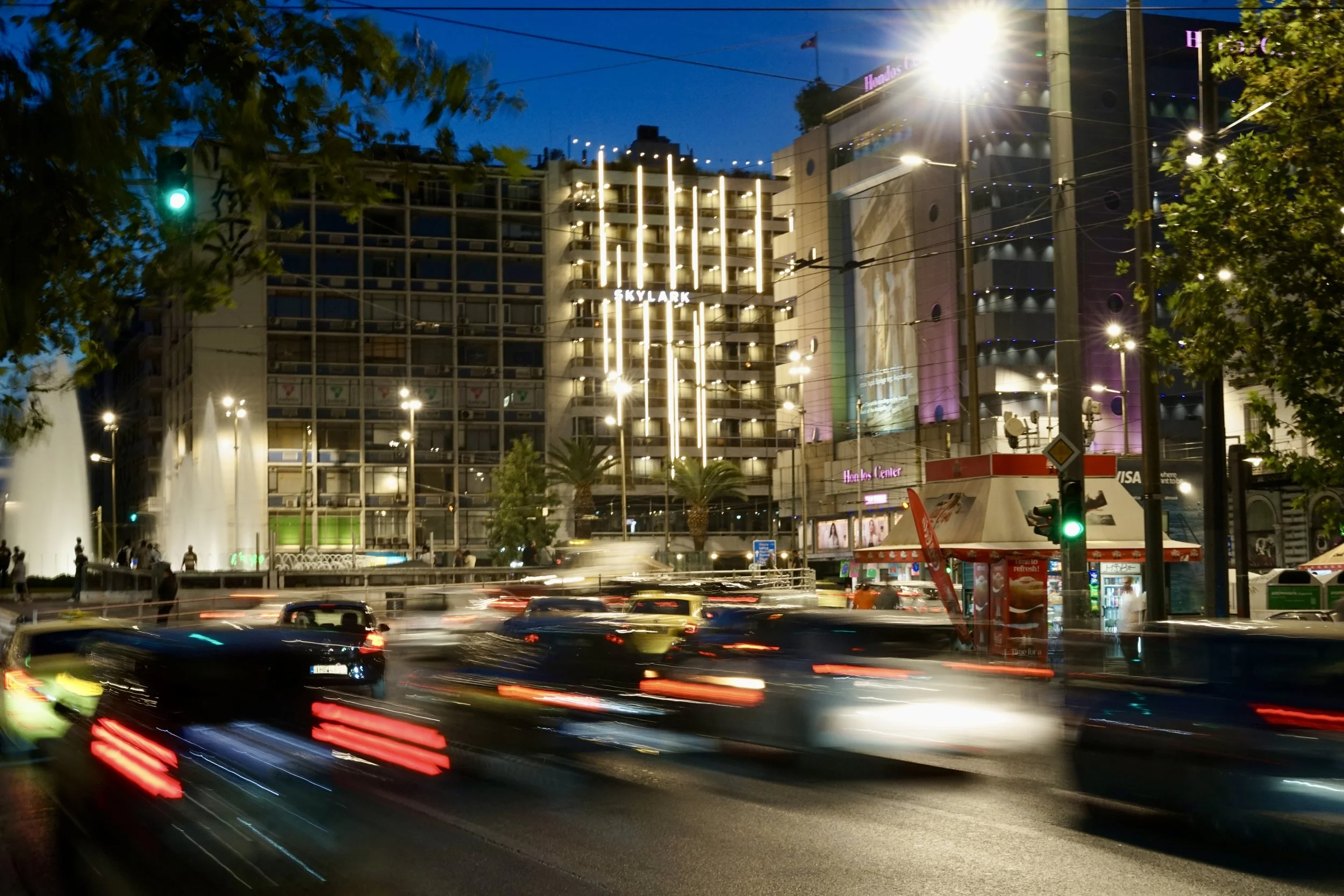 Night traffic light trails blurring past illuminated buildings at Omonoia Square.