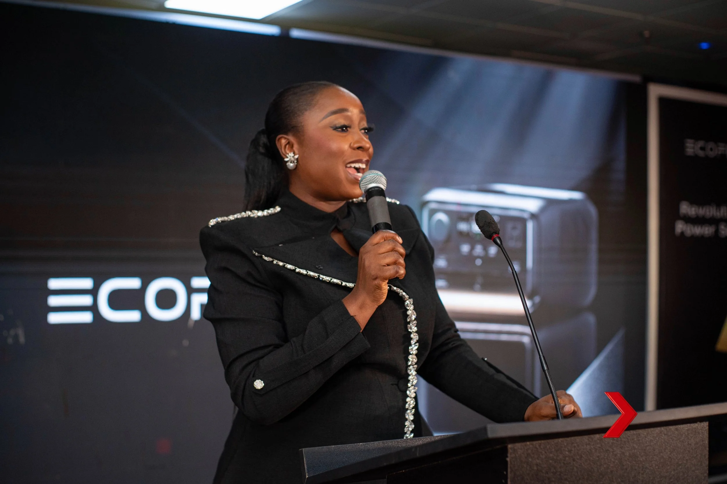 A woman speaking into a microphone at a conference or event, wearing a black blazer with decorative silver accents, standing behind a podium with a red arrow symbol, with a large screen displaying the word 'ECO' in the background.