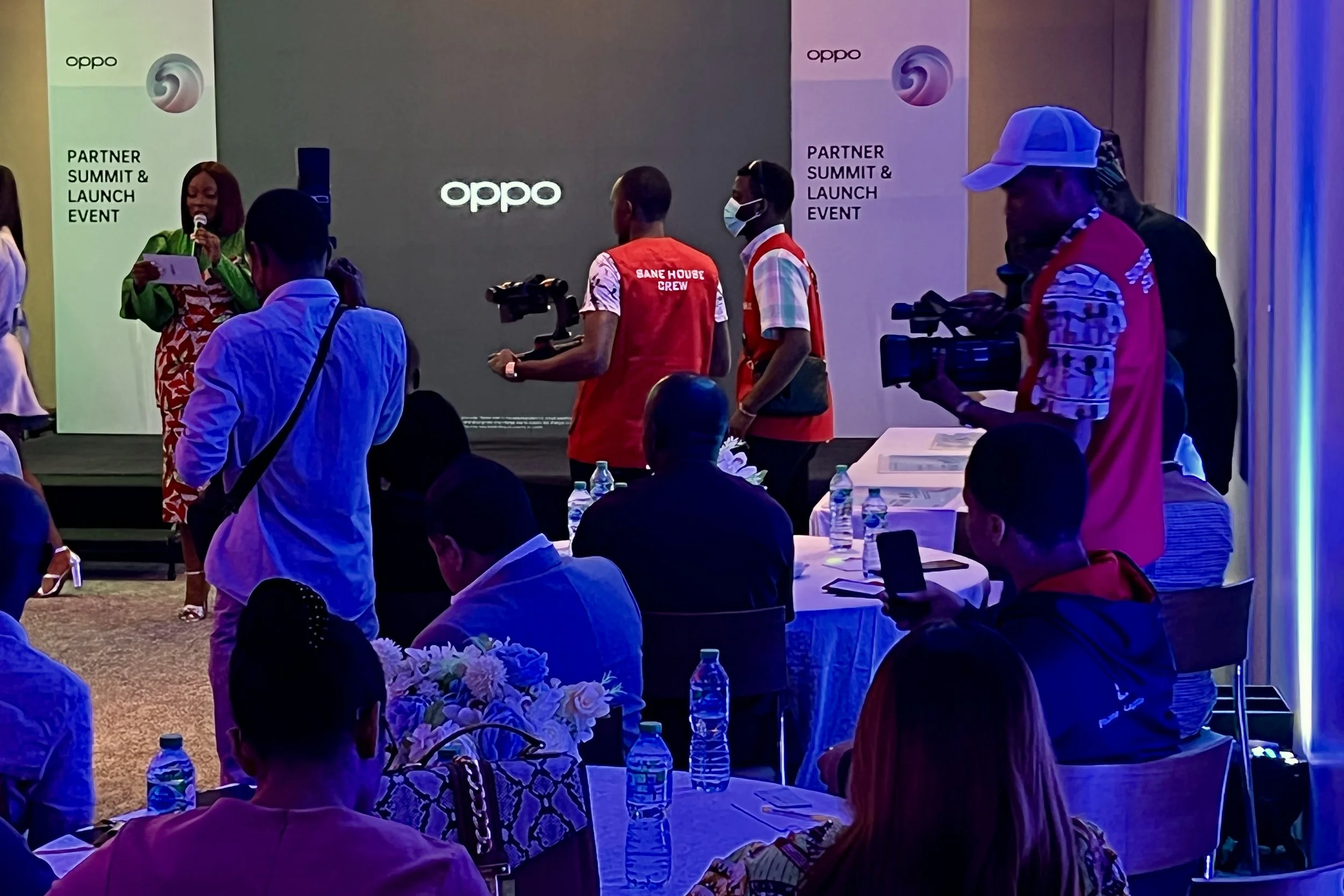 People attending a partner summit and launch event for Oppo, with some wearing red vests and masks, a woman speaking on stage, banners displaying Oppo branding, and water bottles on tables.