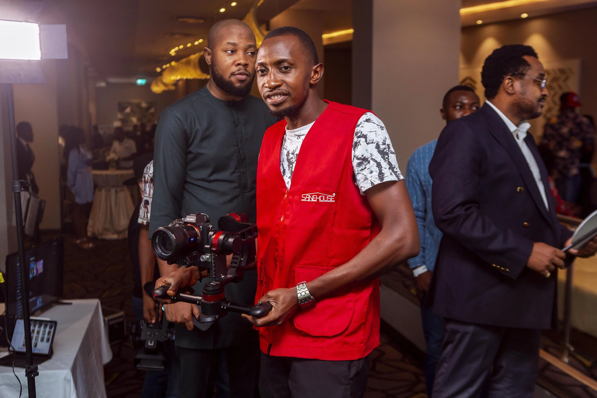 Two men, one wearing a red vest and holding a camera stabilizer, stand close together at an indoor event, with other people in the background.
