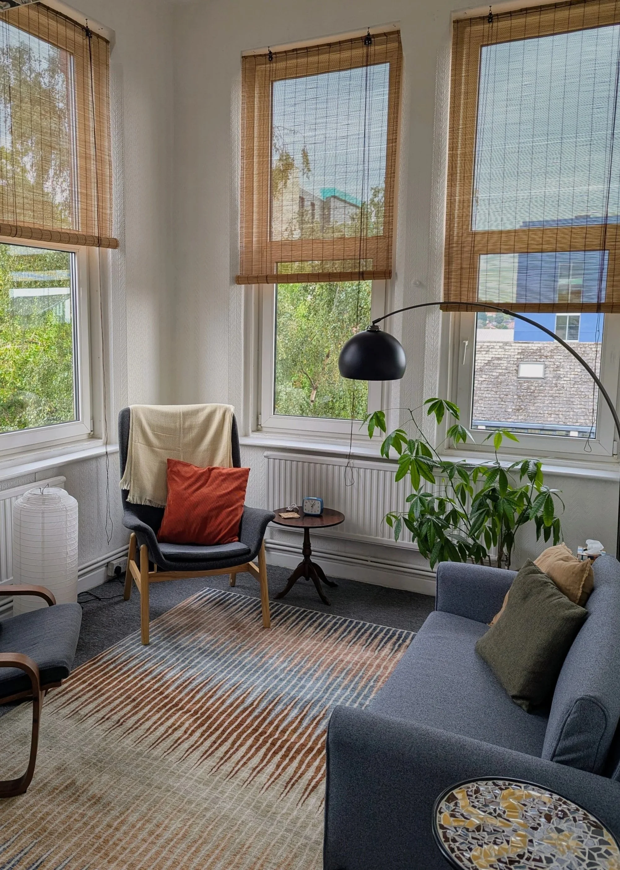 Therapy room with large windows covered by bamboo blinds, two grey armchairs, a small round wooden table, a blue sofa with cushions, a potted plant, a striped area rug, and a black arched floor lamp.