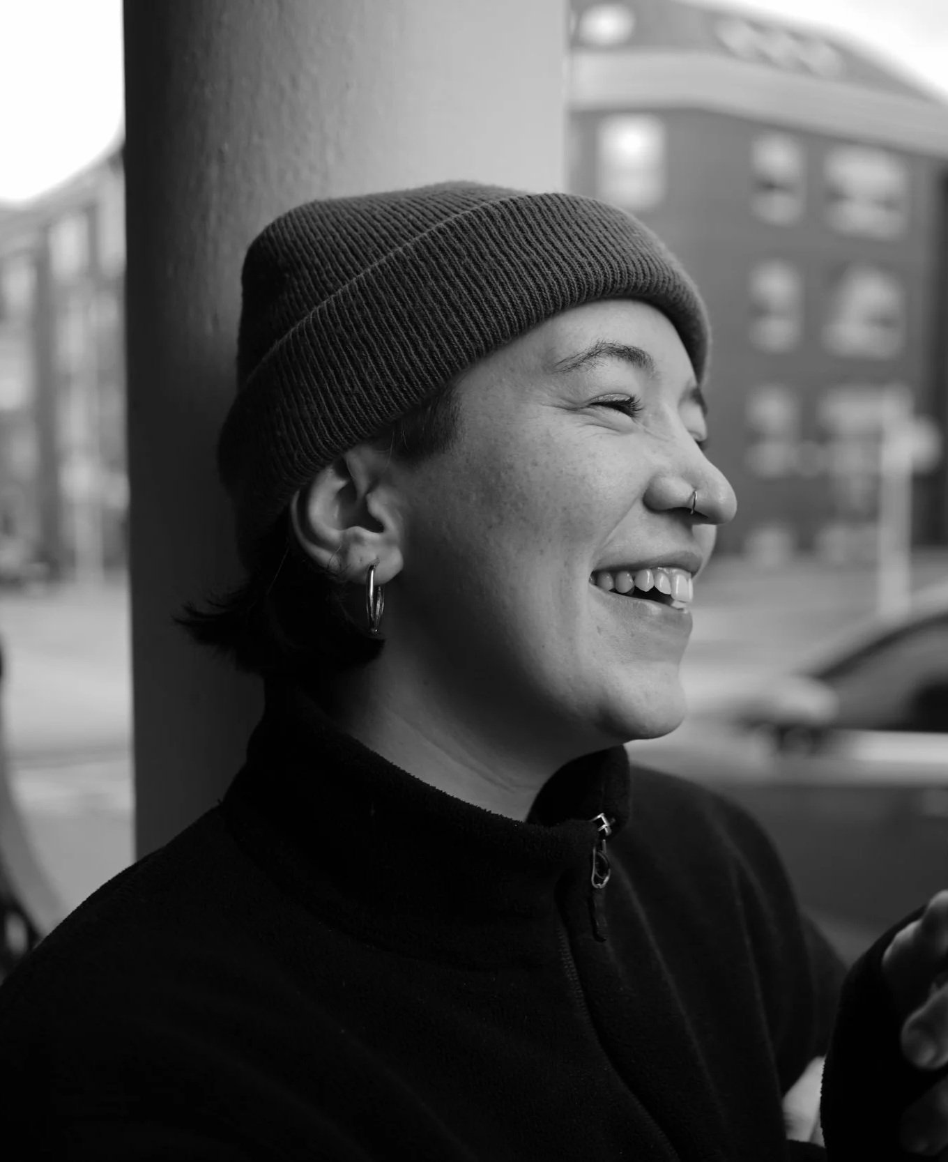 A smiling gender-fluid person with short hair and piercings, wearing a beanie and a black fleece, leaning against a wall in an urban setting.