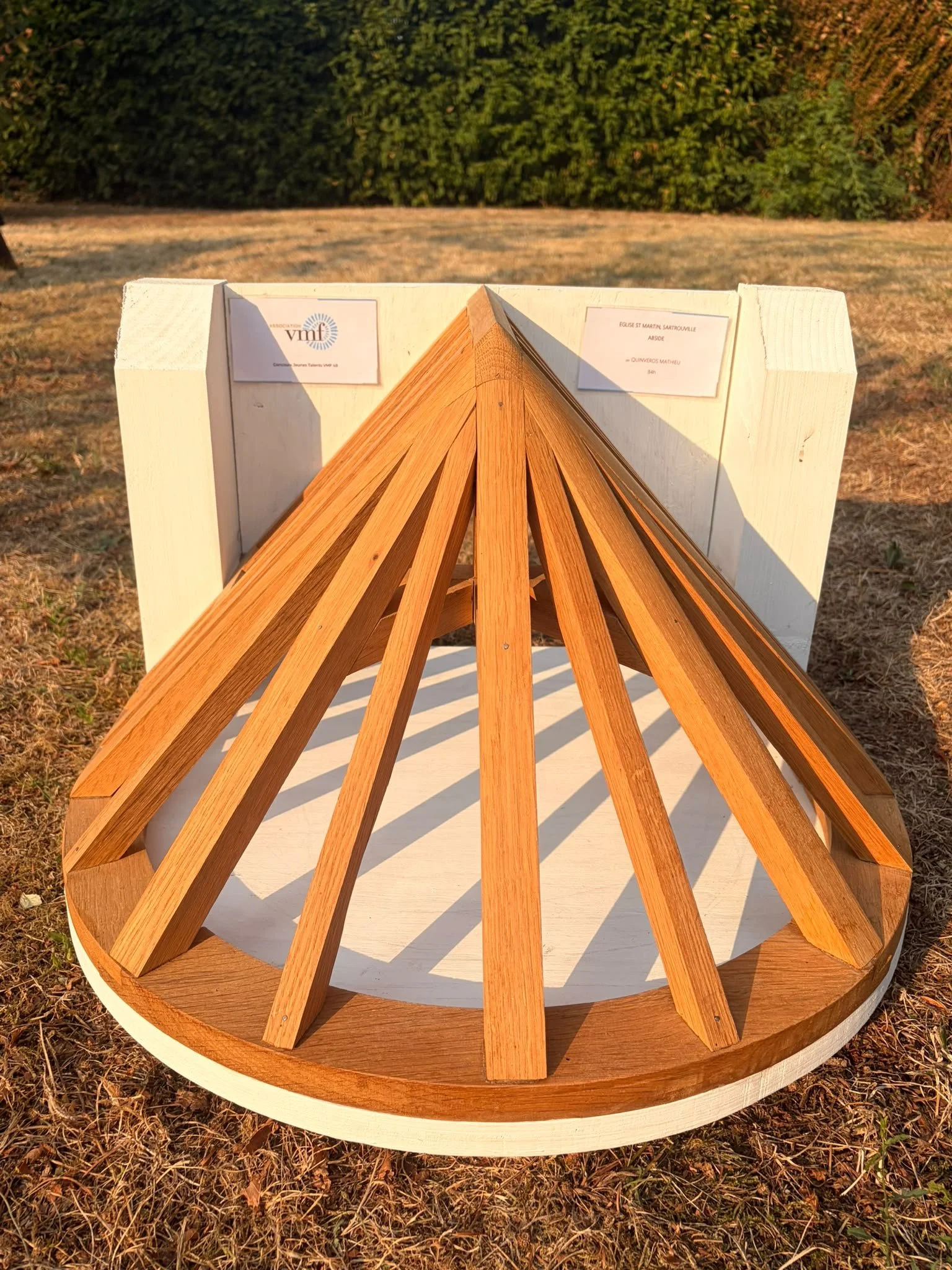 Un modèle de pyramide en bois posé sur une plateforme ronde en bois, avec des étiquettes sur le haut, en extérieur, au coucher de soleil.