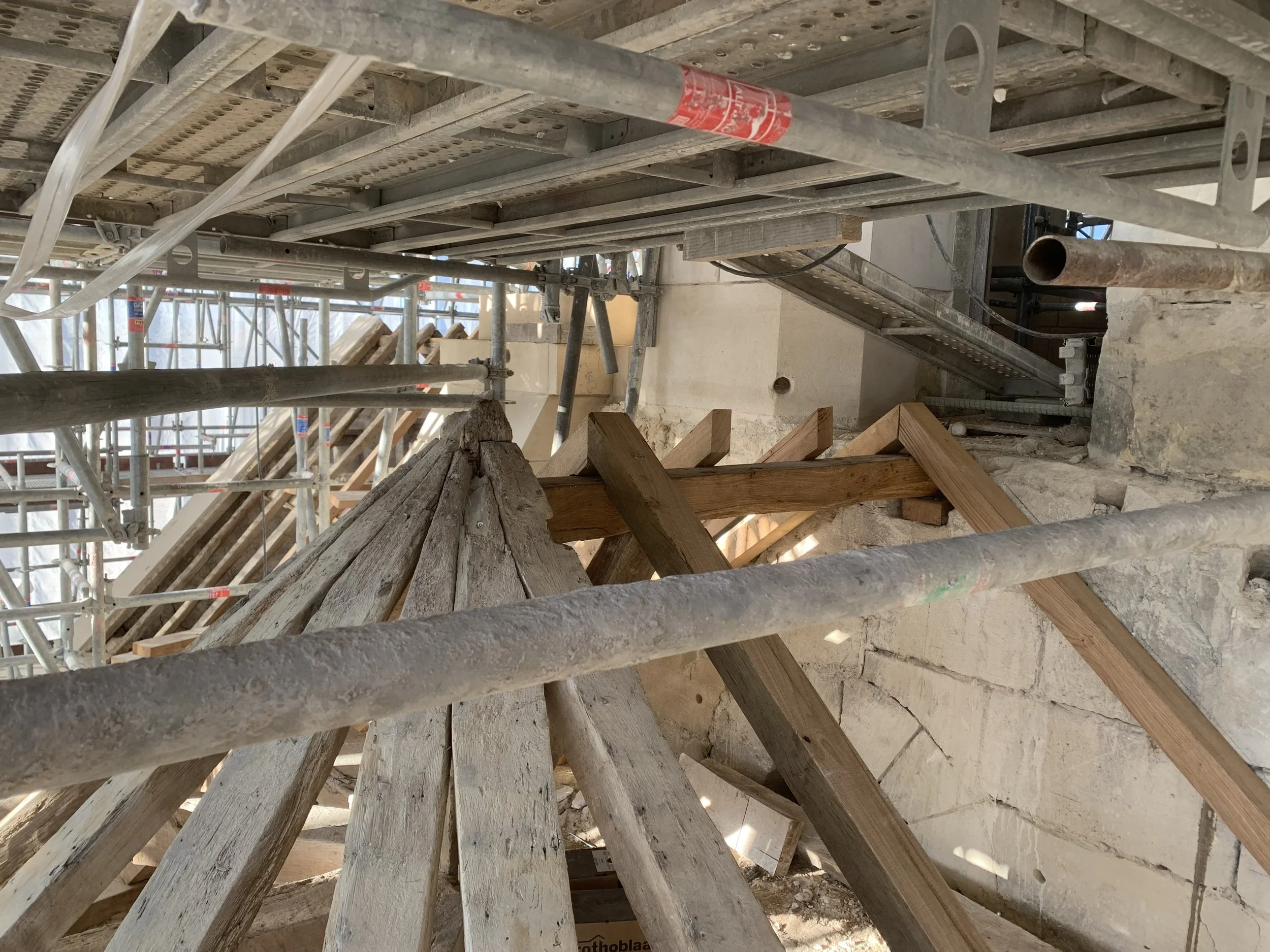Échafaudage en métal sur un chantier de construction avec des éléments en bois, un sol en béton, et plusieurs tuyaux visibles.